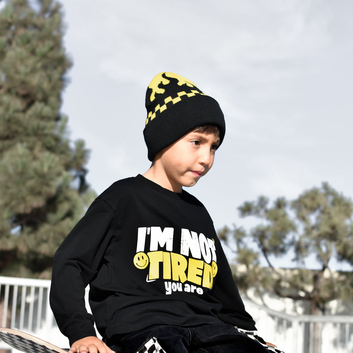 A child wearing a black graphic sweatshirt with yellow and white text, a black beanie with yellow checkers, and black pants with a checkered pattern, sitting on a skateboard outdoors.