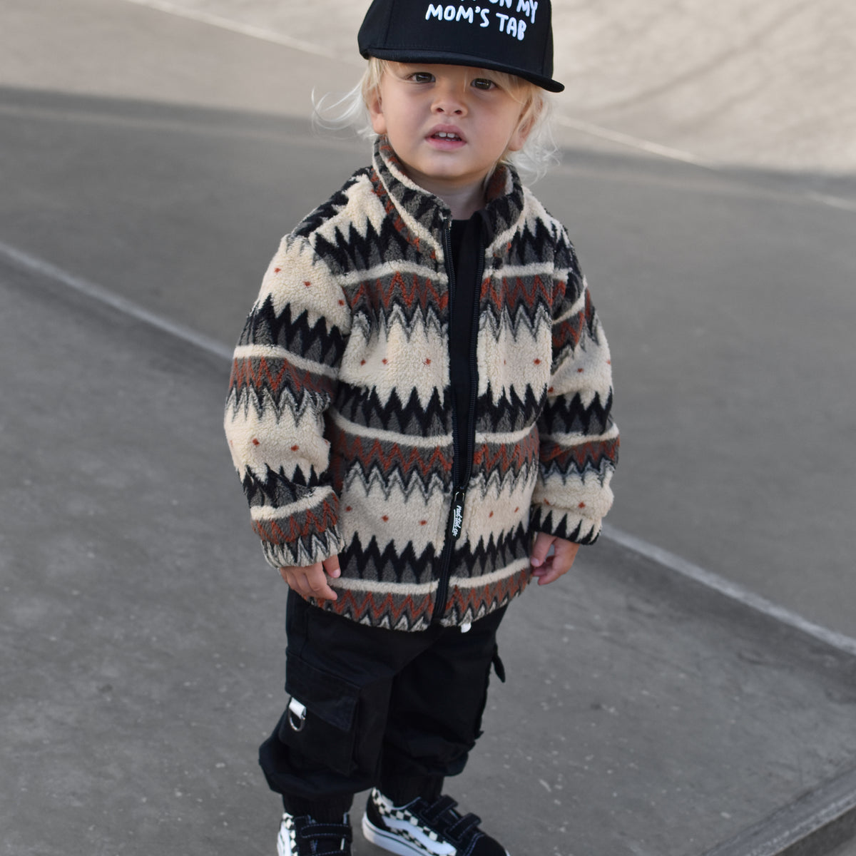 A toddler standing outdoors on concrete steps, wearing a cream and brown geometric patterned zip-up jacket, black pants, black sneakers, and a black cap with white text that reads 'PUT IT ON MY MOM'S TAB'.