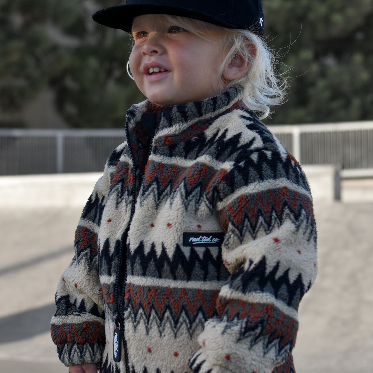 A toddler wearing a cream, black, and rust-colored geometric patterned fleece jacket and a black baseball cap that says put it on my moms tab, standing outdoors at a skatepark.