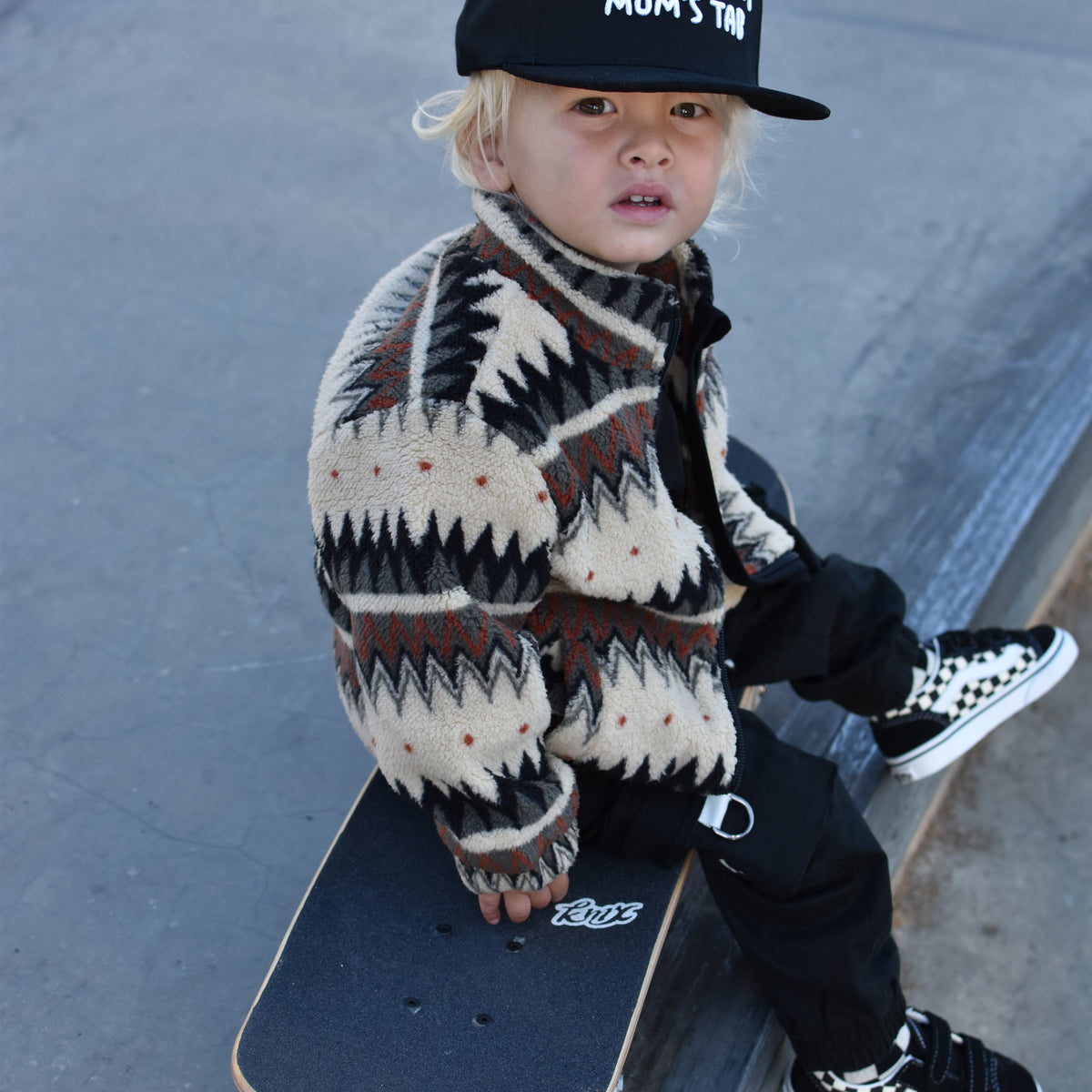 A toddler sits on a skateboard on a concrete ramp, wearing a black cap with white text, a cream and black patterned knit sweater, black pants, and black-and-white checkered sneakers.