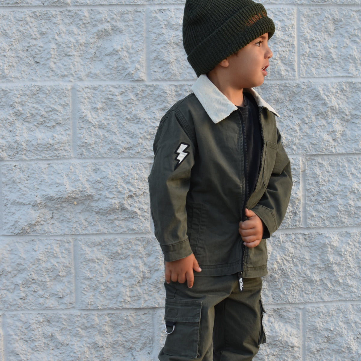 A young child wearing an olive green two-piece jacket and cargo pants set with a white collar, a black beanie, and black sneakers, standing against a light gray textured brick wall.