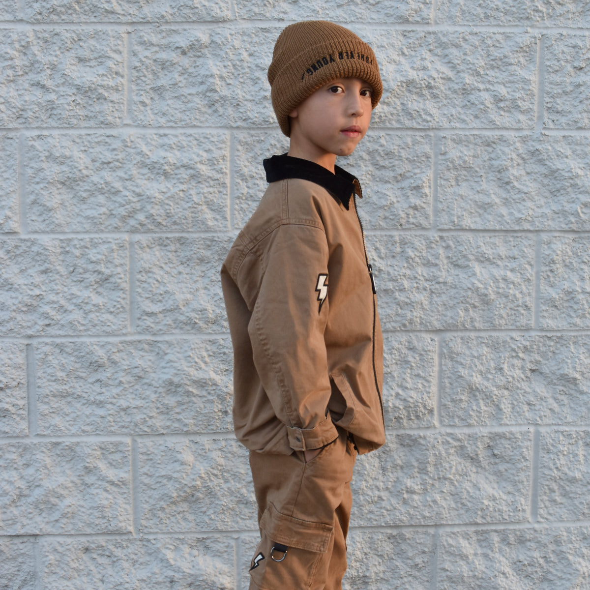 A child wearing a brown beanie with 'Forever Young' text, a matching brown jacket and cargo pants, and white sneakers, standing against a light gray textured brick wall.
