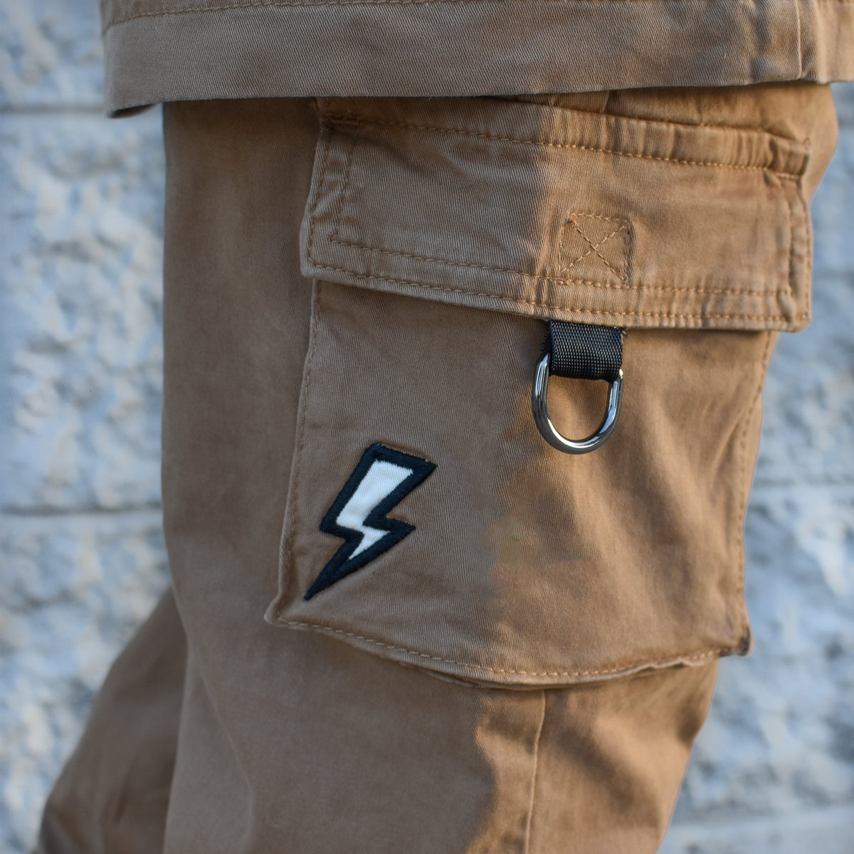 Close-up of tan cargo pants worn by a person, showing a flap pocket with a black-and-white lightning bolt patch and a metal ring detail, against a light textured wall.