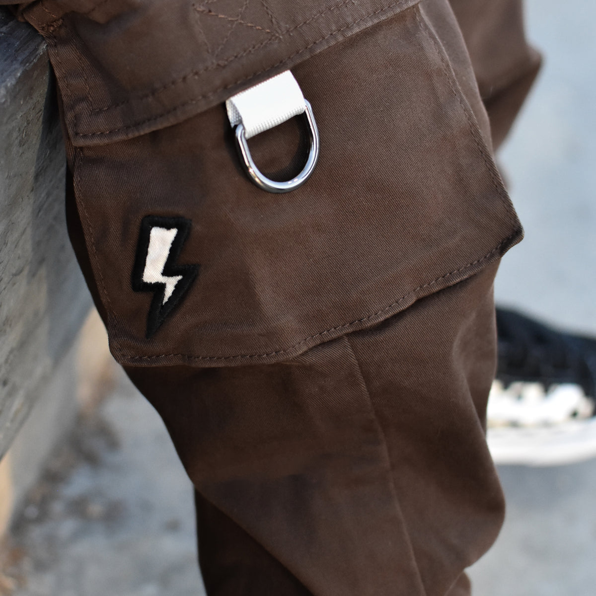 Close-up of brown cargo pants featuring a silver metal D-ring on the cargo pocket and a black-and-white lightning bolt patch.