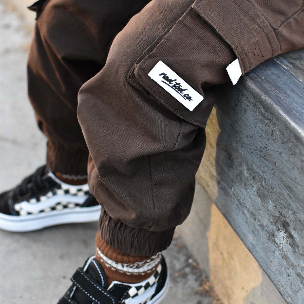 A close-up lifestyle shot of a child wearing brown cargo jogger pants with a visible rad tod co brand patch, paired with black and white sneakers, sitting on a concrete ledge.