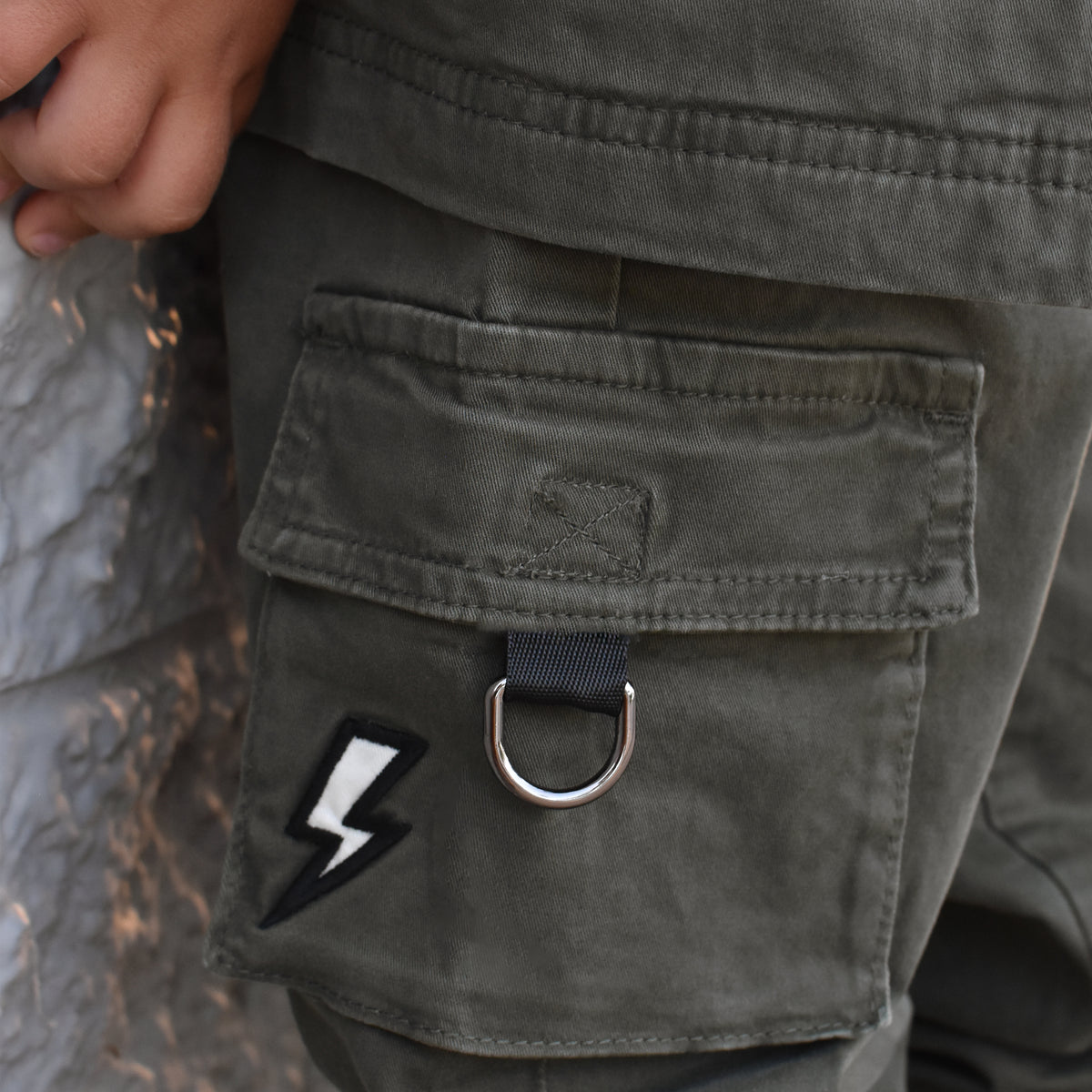 Close-up of a person wearing olive green cargo pants, showing a large front flap pocket with a silver metal D-ring and a black-and-white lightning bolt patch.