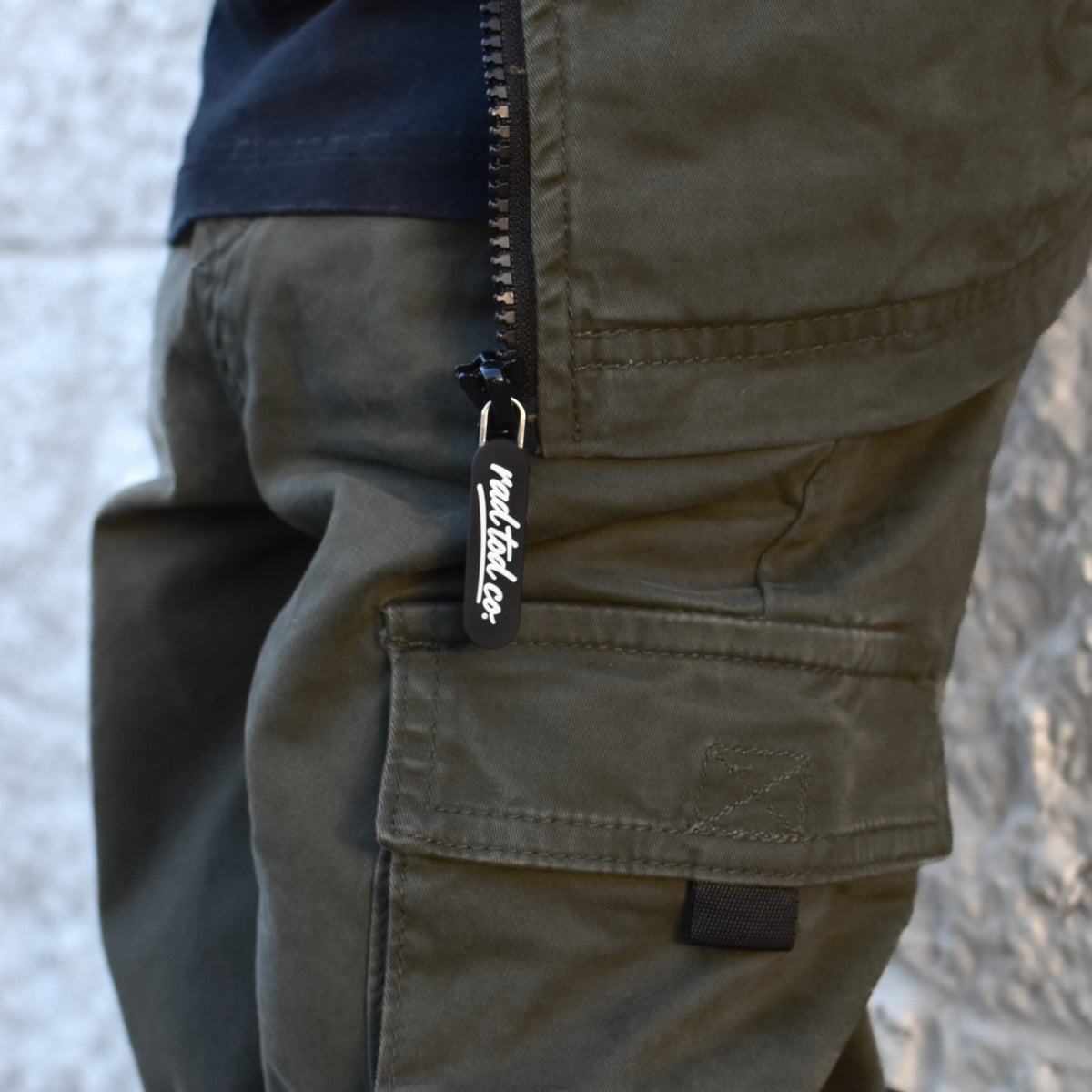Close-up of olive green cargo pants with a black zipper and a visible 'rad tod co.' brand tab, worn with a black shirt against a light textured wall.