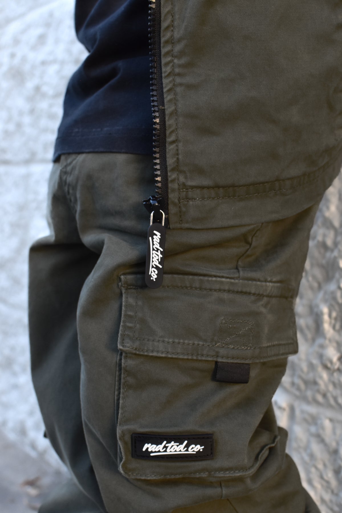 Close-up of olive green cargo pants with a black zipper and a visible 'rad tod co.' brand tab, worn with a black shirt against a light textured wall.