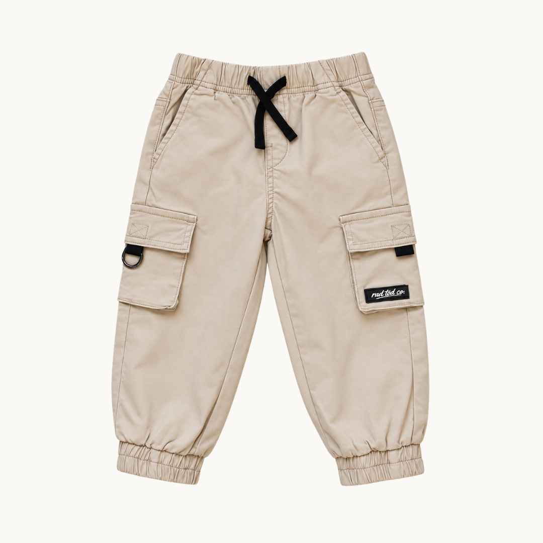 A pair of beige cargo pants for kids, featuring elastic waistband with a black drawstring, side cargo pockets, and elastic cuffs. A small black brand patch is visible on the right pocket flap.