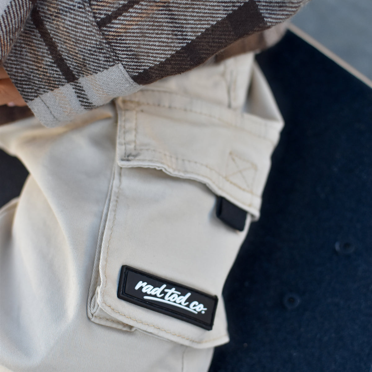 Close-up of a person wearing off-white cargo pants with a black patch that reads 'rad tod co.' and a brown, gray, and white plaid shirt, resting on a dark blue surface.