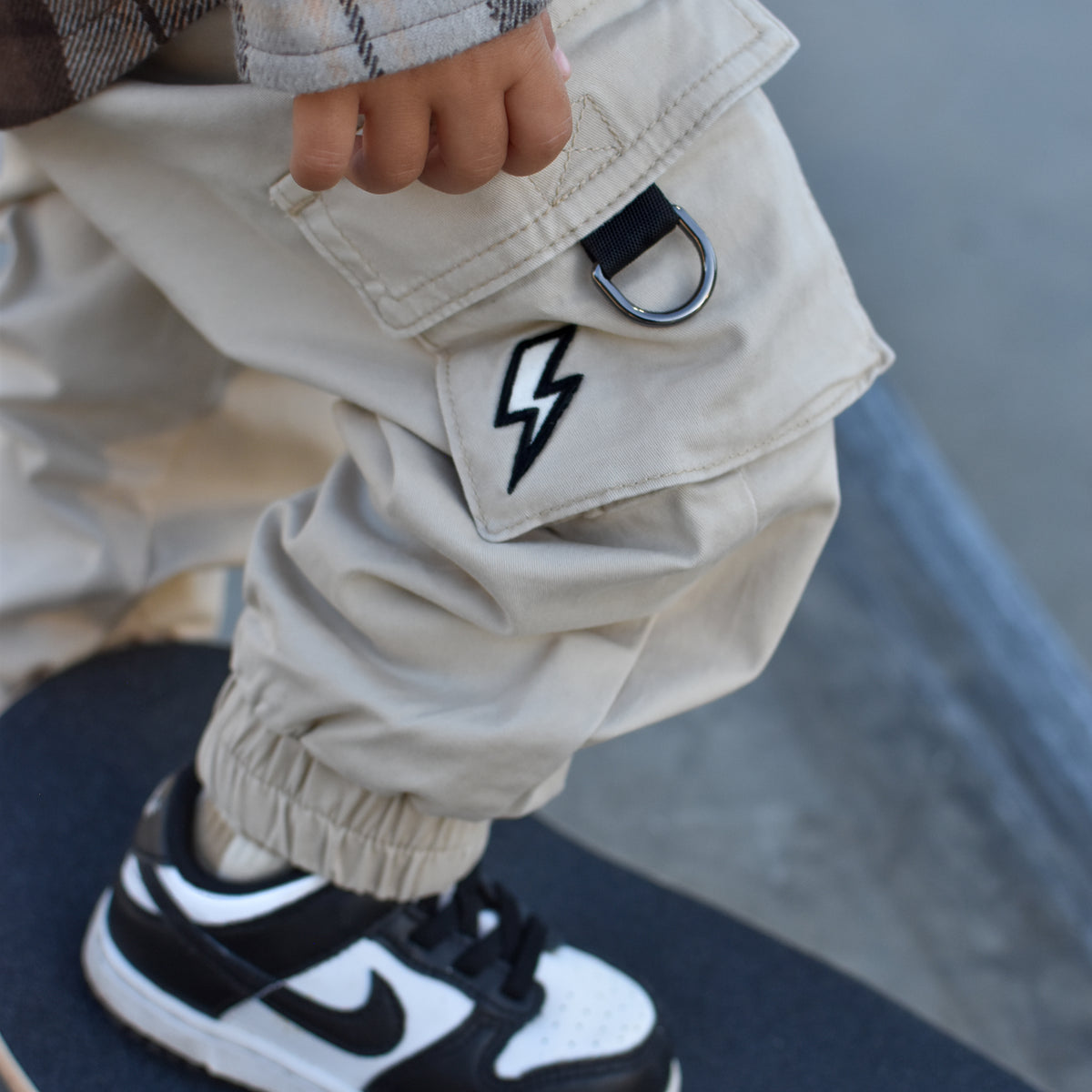 A close-up lifestyle shot of a child wearing beige cargo pants with a black lightning-bolt patch on the pocket, a brown plaid shirt, and a black and white Nike sneaker while standing on a skateboard.