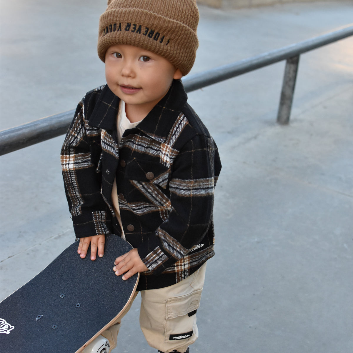 A toddler standing at a skatepark holding a skateboard, wearing a brown knit beanie with black text, a black and brown plaid flannel shirt, beige cargo pants, and black and white sneakers.
