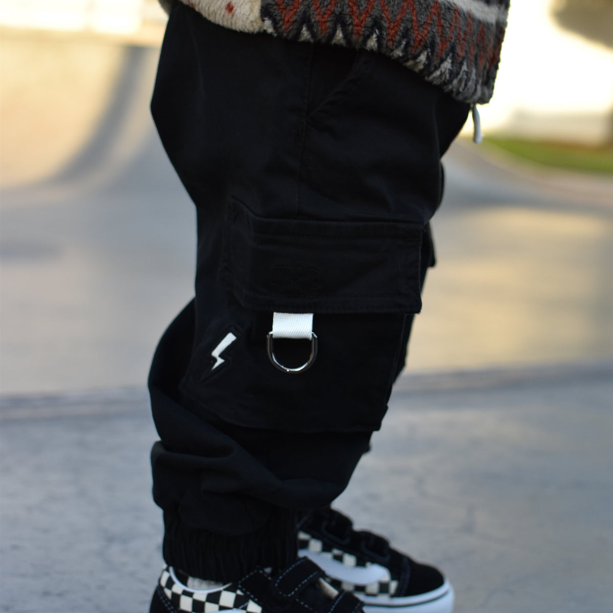 A close-up of a child's legs wearing black cargo pants with a silver D-ring detail, a patterned knit sweater, and black-and-white checkered Vans sneakers on a paved outdoor surface.