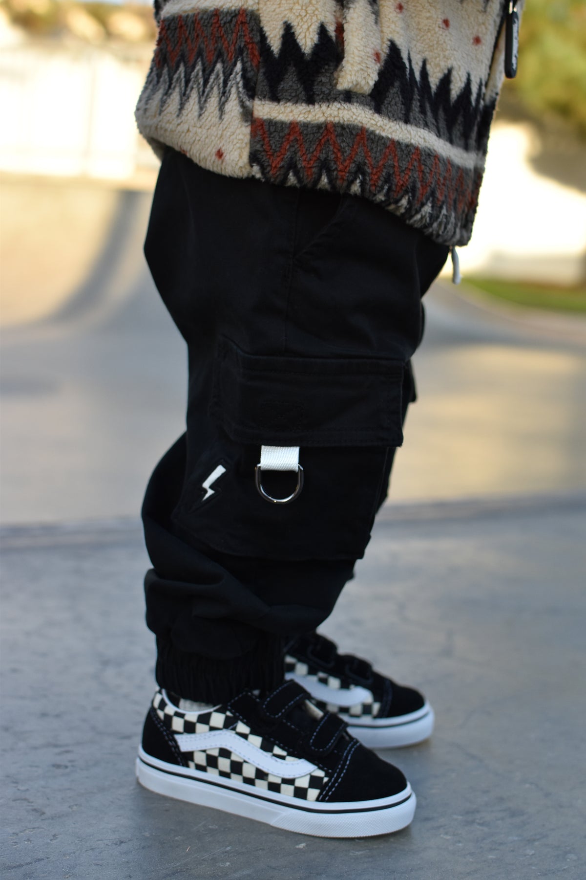 A close-up of a child's legs wearing black cargo pants with a silver D-ring detail, a patterned knit sweater, and black-and-white checkered Vans sneakers on a paved outdoor surface.