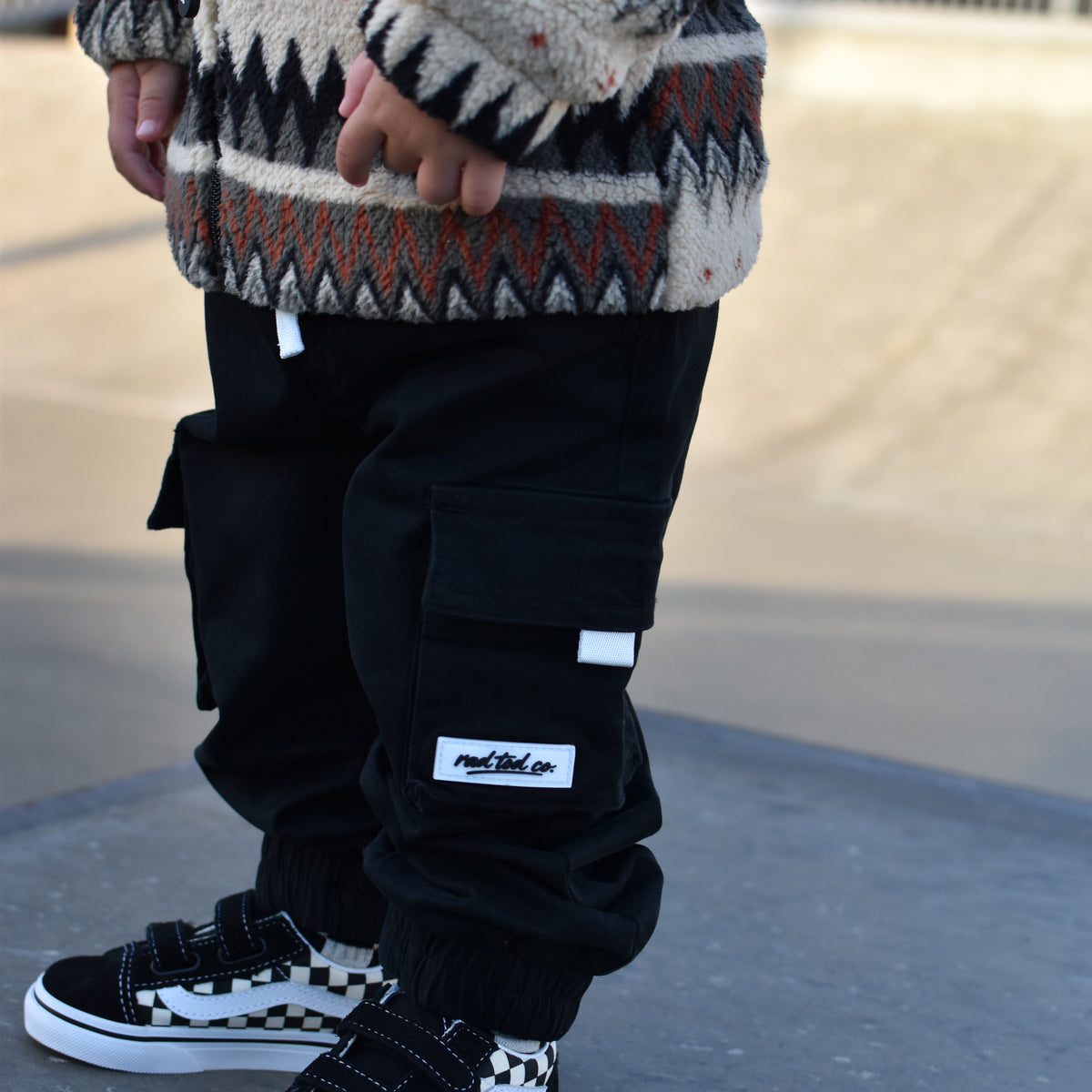 A child wearing black cargo pants with a white 'rad tod co' brand patch, paired with a patterned knit sweater and black-and-white checkered sneakers, standing outdoors on a concrete surface.