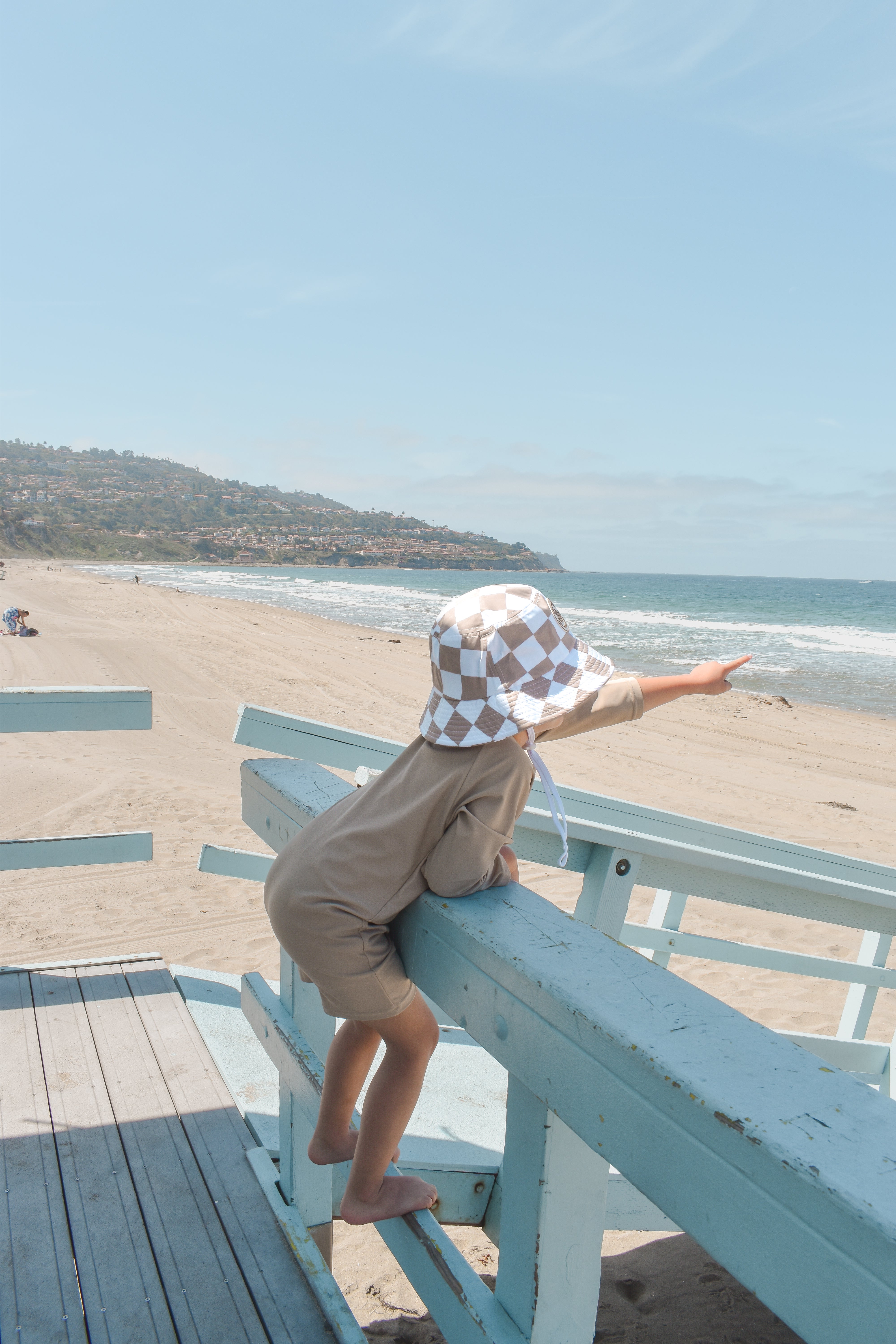 Young boy standing on a lifeguard tower at the beach, wearing a tan one-piece rashguard with a front zipper and enjoying the view