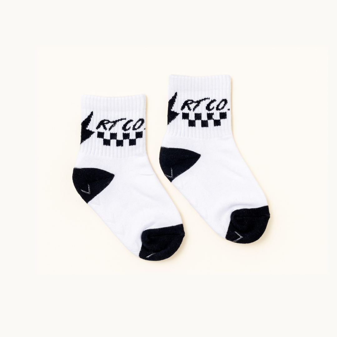A pair of white crew socks with black accents, featuring a black and white checkered pattern and the brand name 'RT CO.' printed on the cuff, displayed on a light cream background.