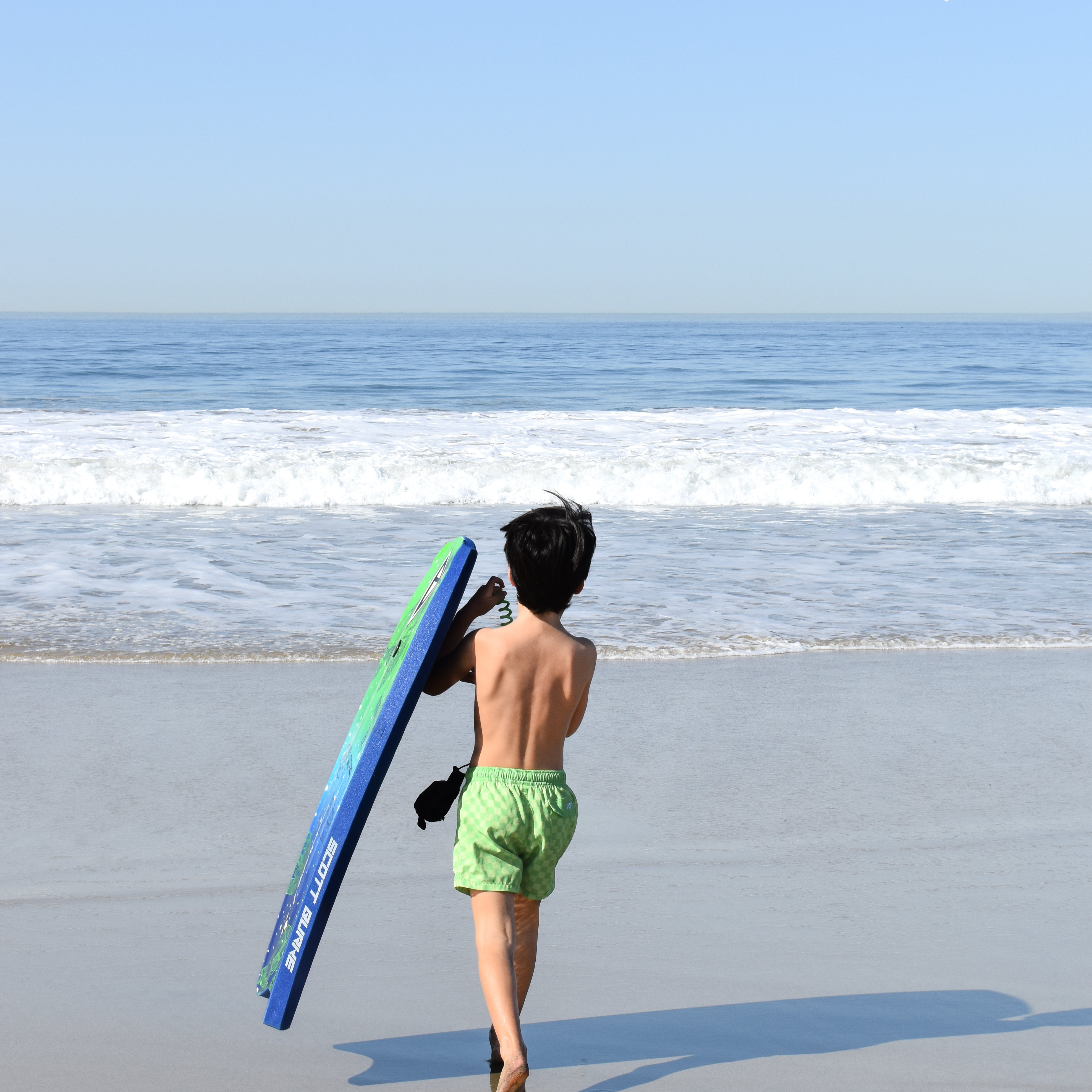 boy walking on a beach with a blue surfboard, wearing lime green shorts by rad toddler