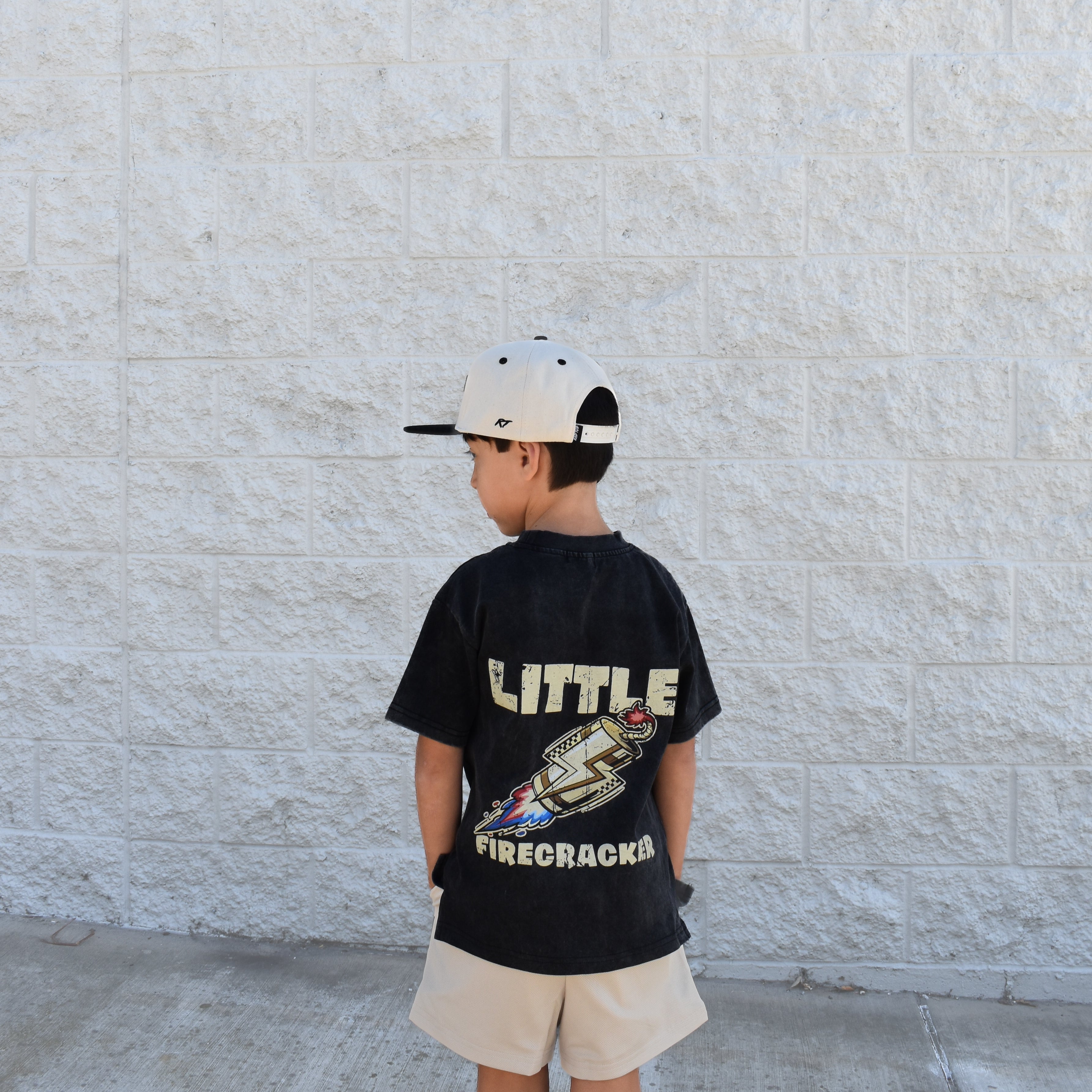 Child wearing a black t-shirt with text and graphics, standing against a light stone wall.