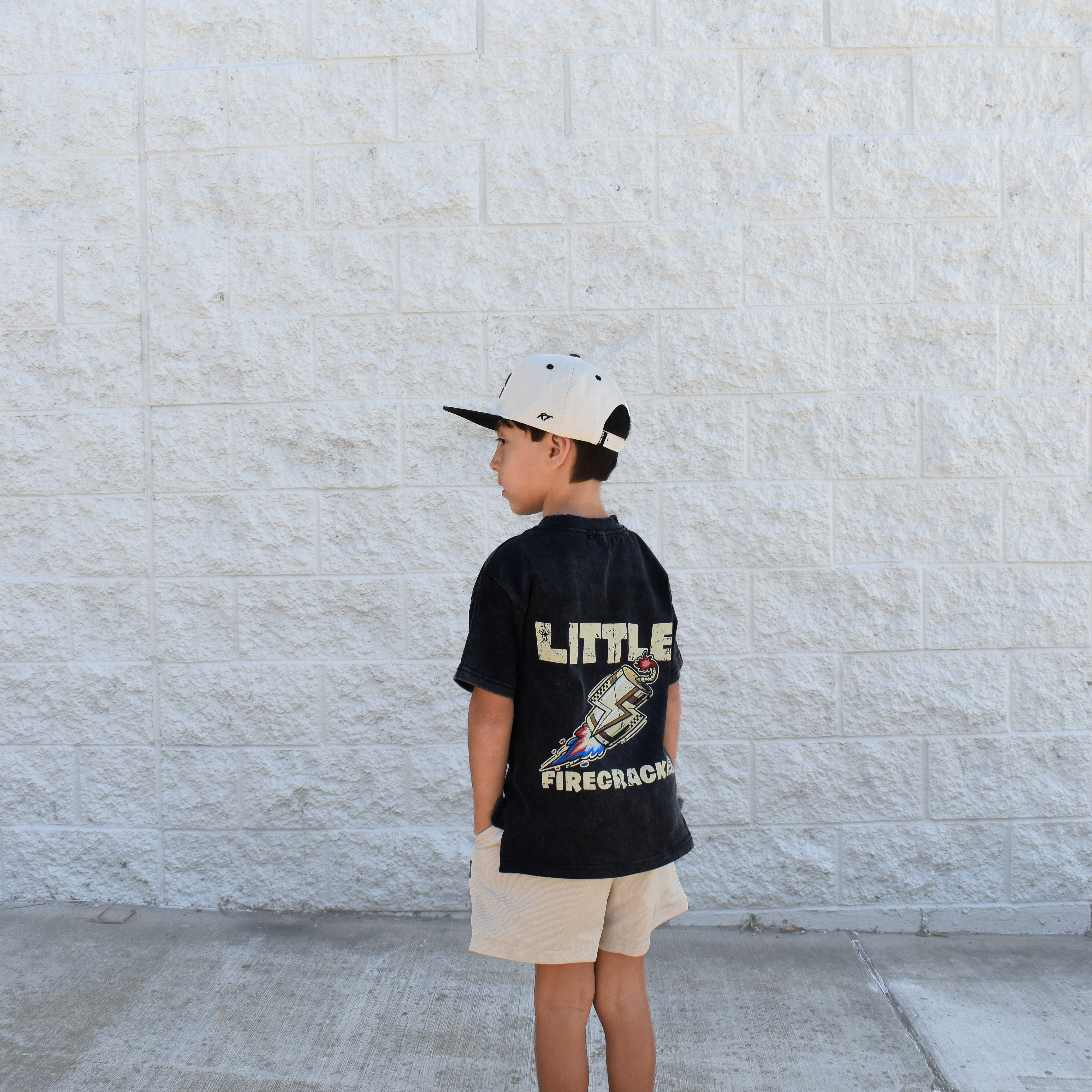 Child wearing a black t-shirt with 'LITTLE FIRECRACKER' text, standing against a light gray wall.