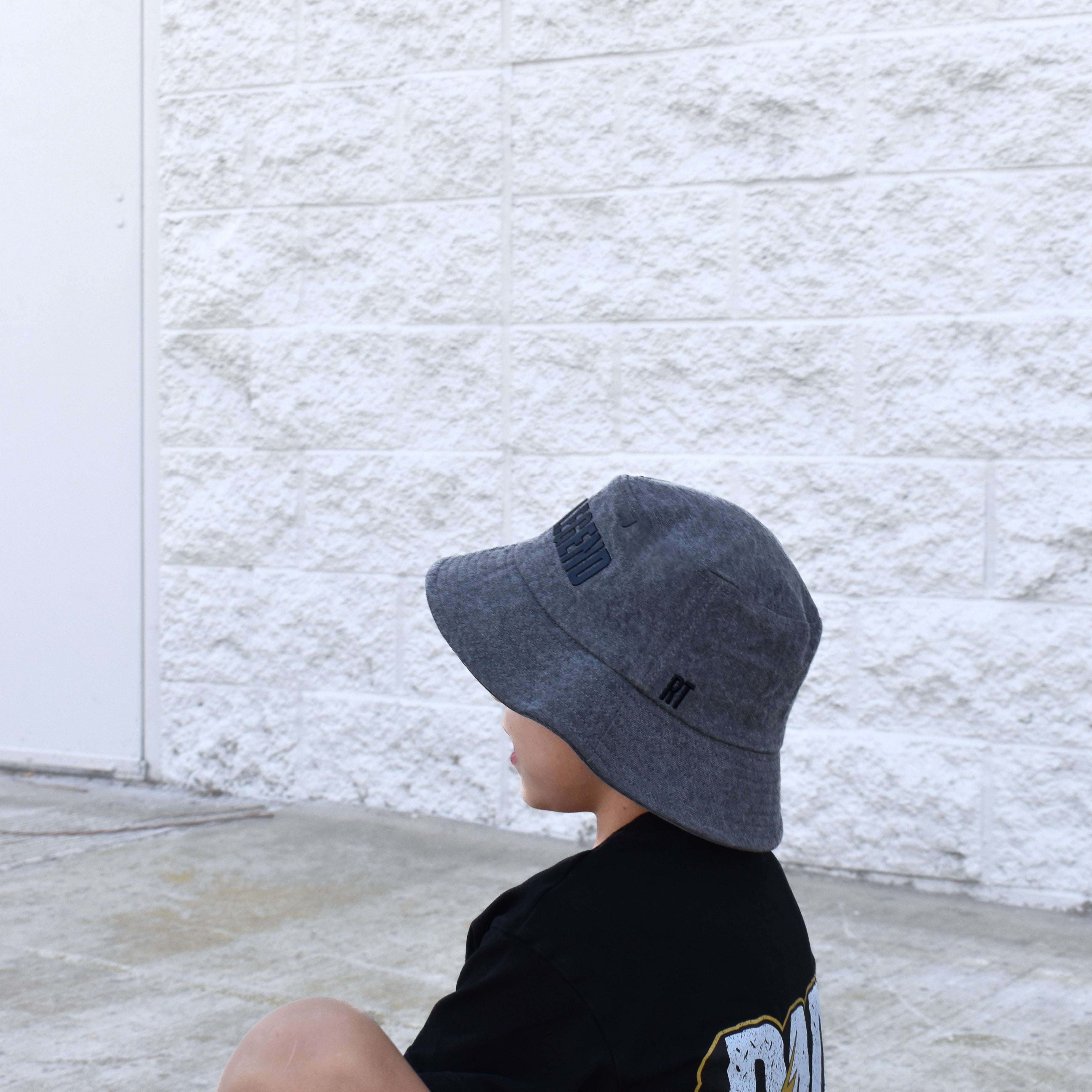 child wearing a gray bucket hat sitting against a textured white wall.