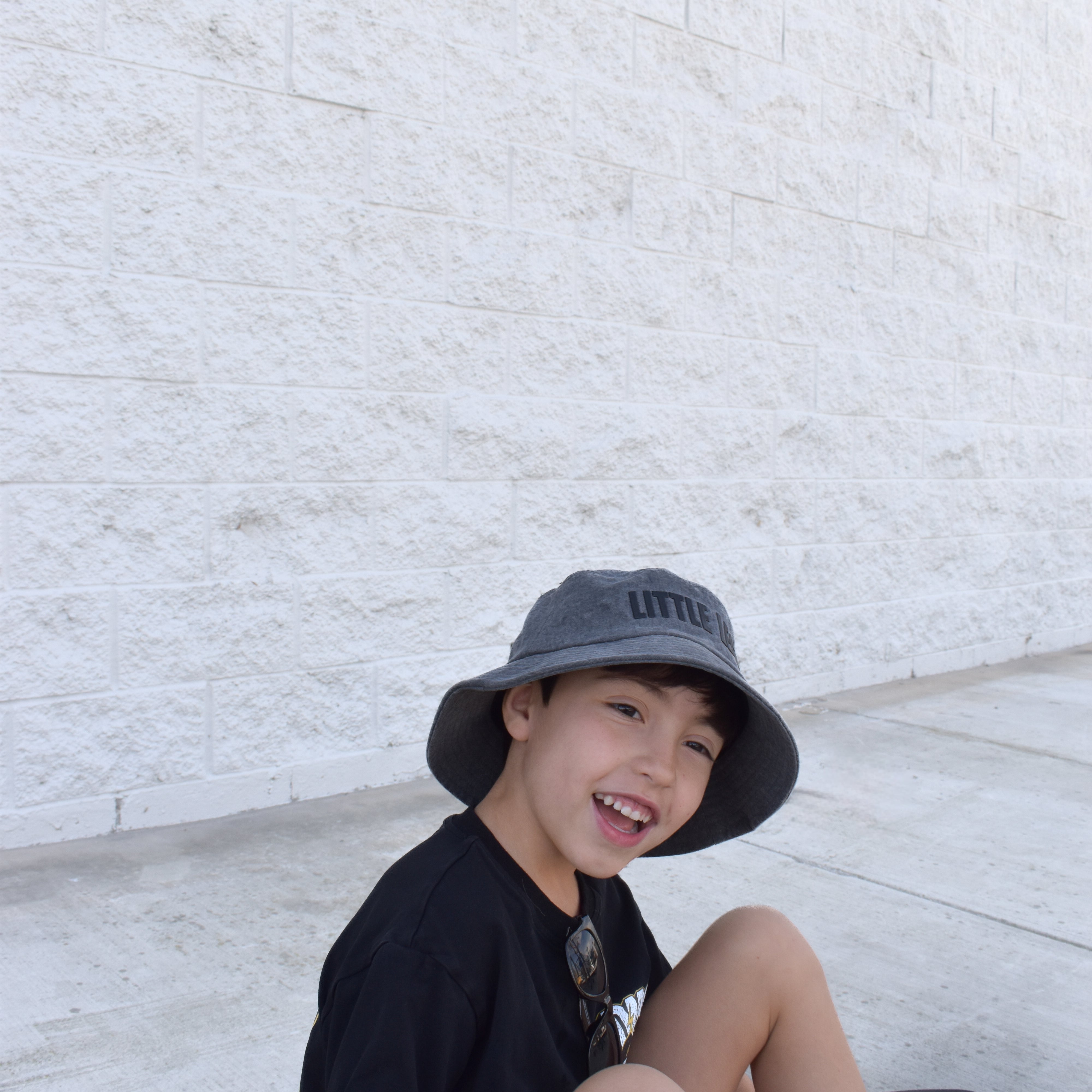 Child sitting on a step wearing a black shirt, gray bucket hat, and white shoes against a light stone wall.