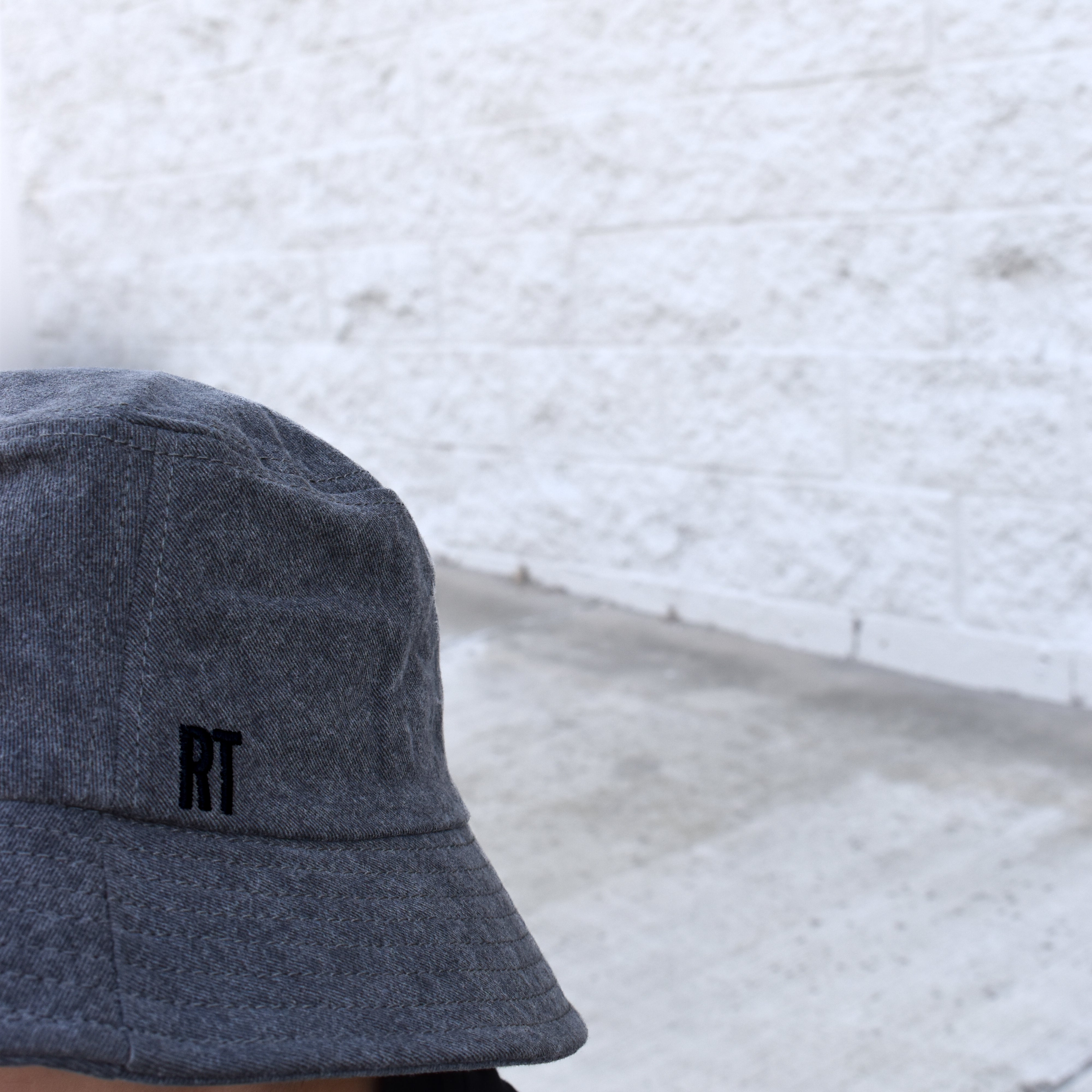 Person wearing a gray bucket hat with 'RT' logo against a light gray textured wall.