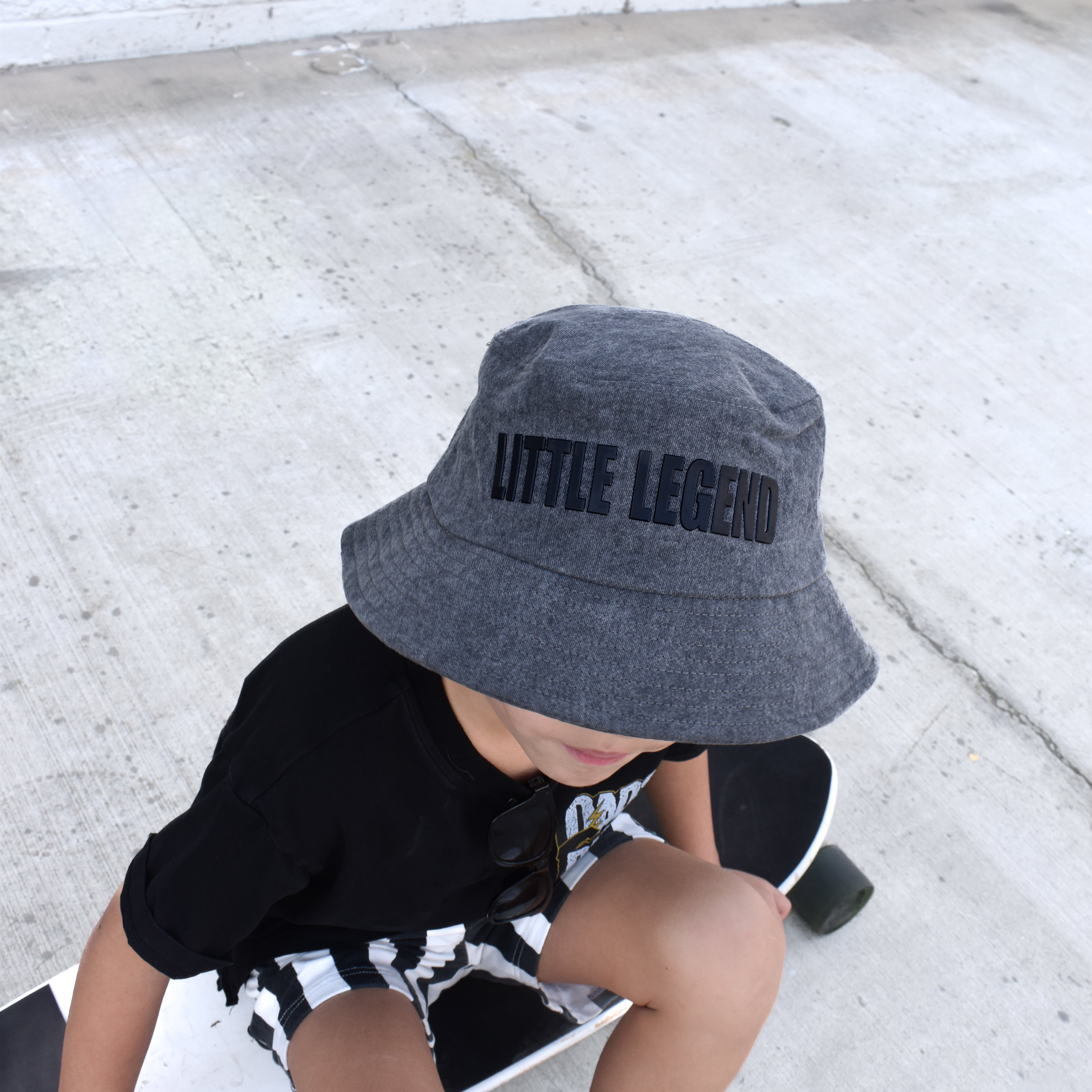 Child wearing a gray bucket hat with 'LITTLE LEGEND' text, sitting on a skateboard.