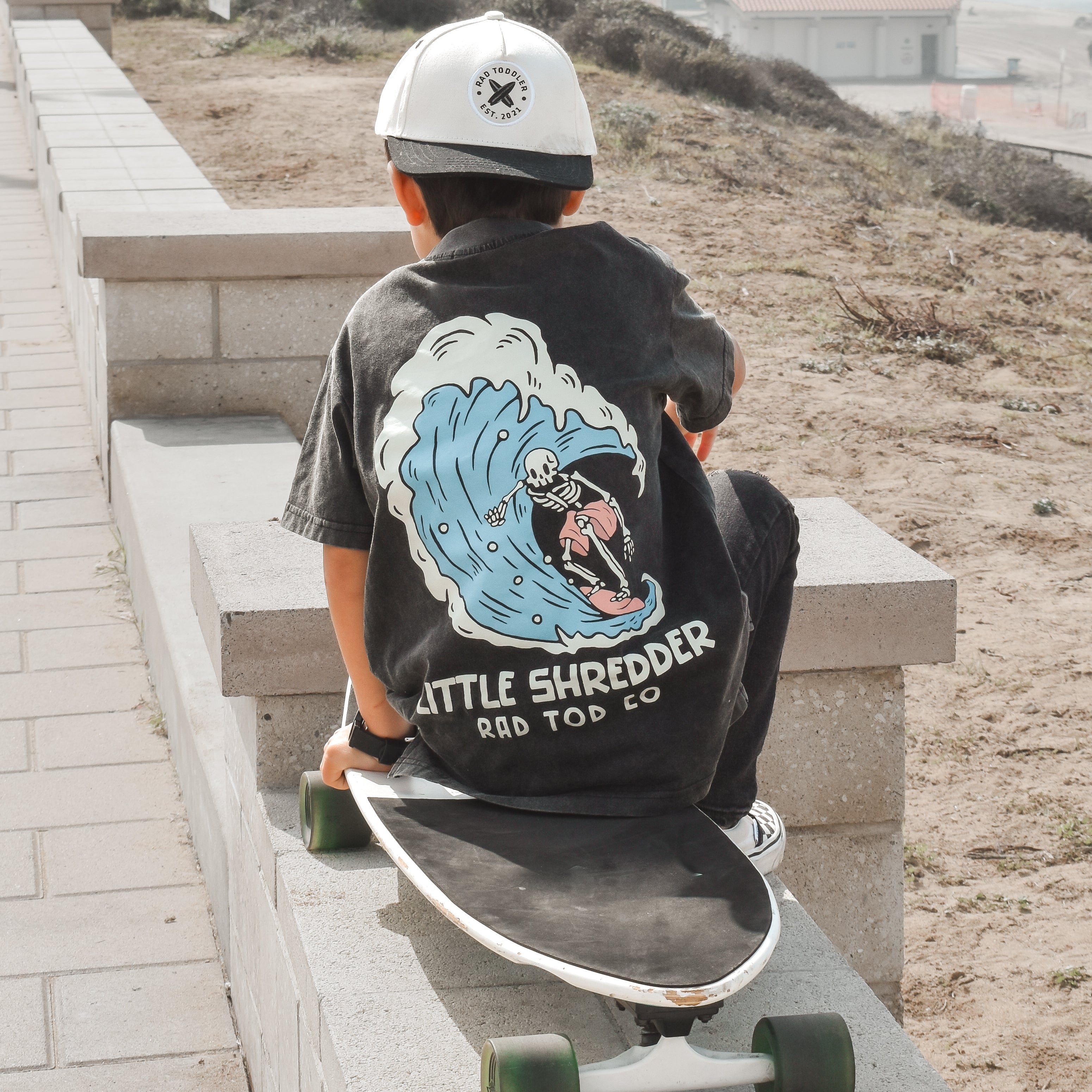 a boy wearing RAD TODS Kids' 'Little Shredder' T-shirt featuring a skeleton surfing a wave design, perfect for young surfers and adventure lovers.