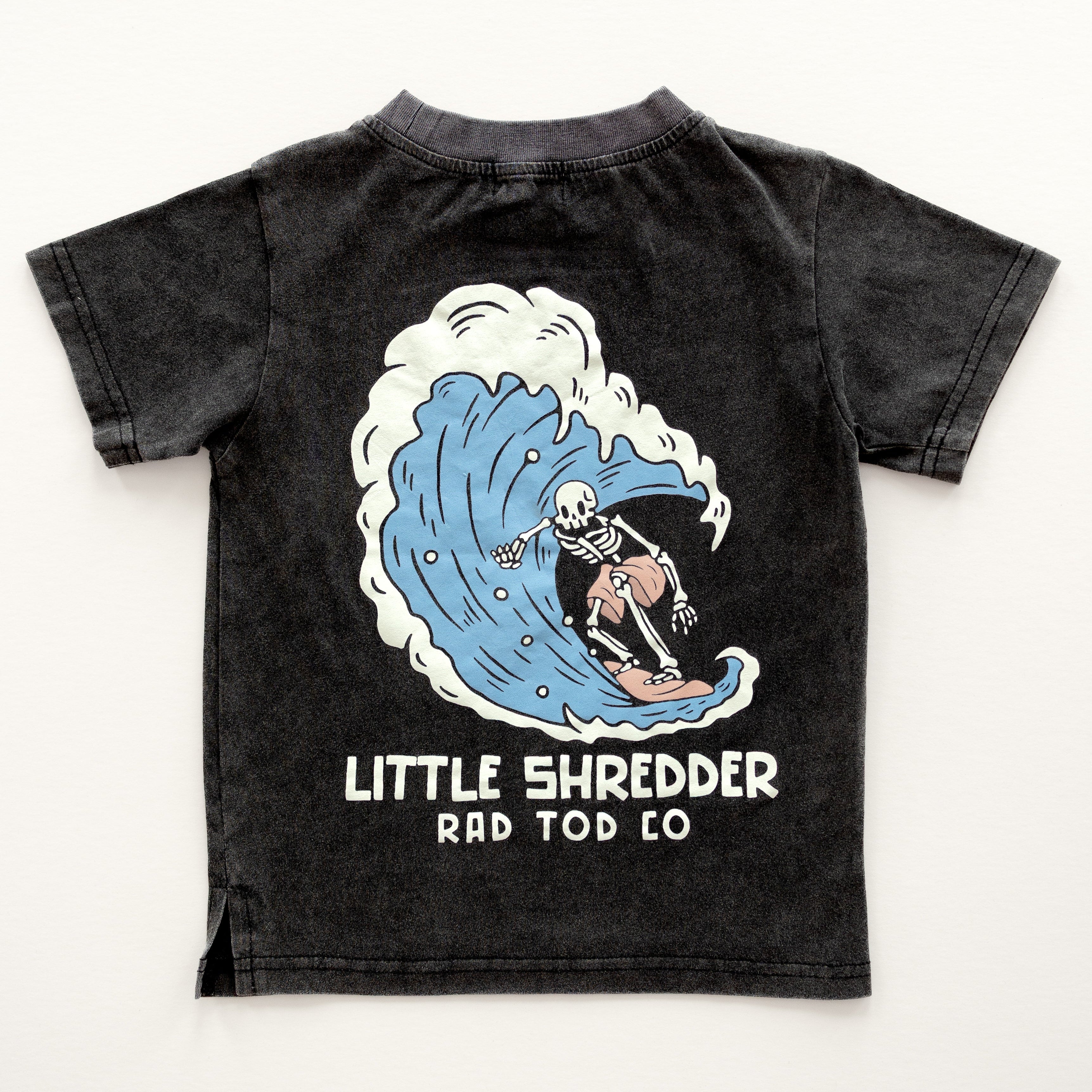 Kids' 'Little Shredder' T-shirt featuring a skeleton surfing a wave design, perfect for young surfers and adventure lovers.