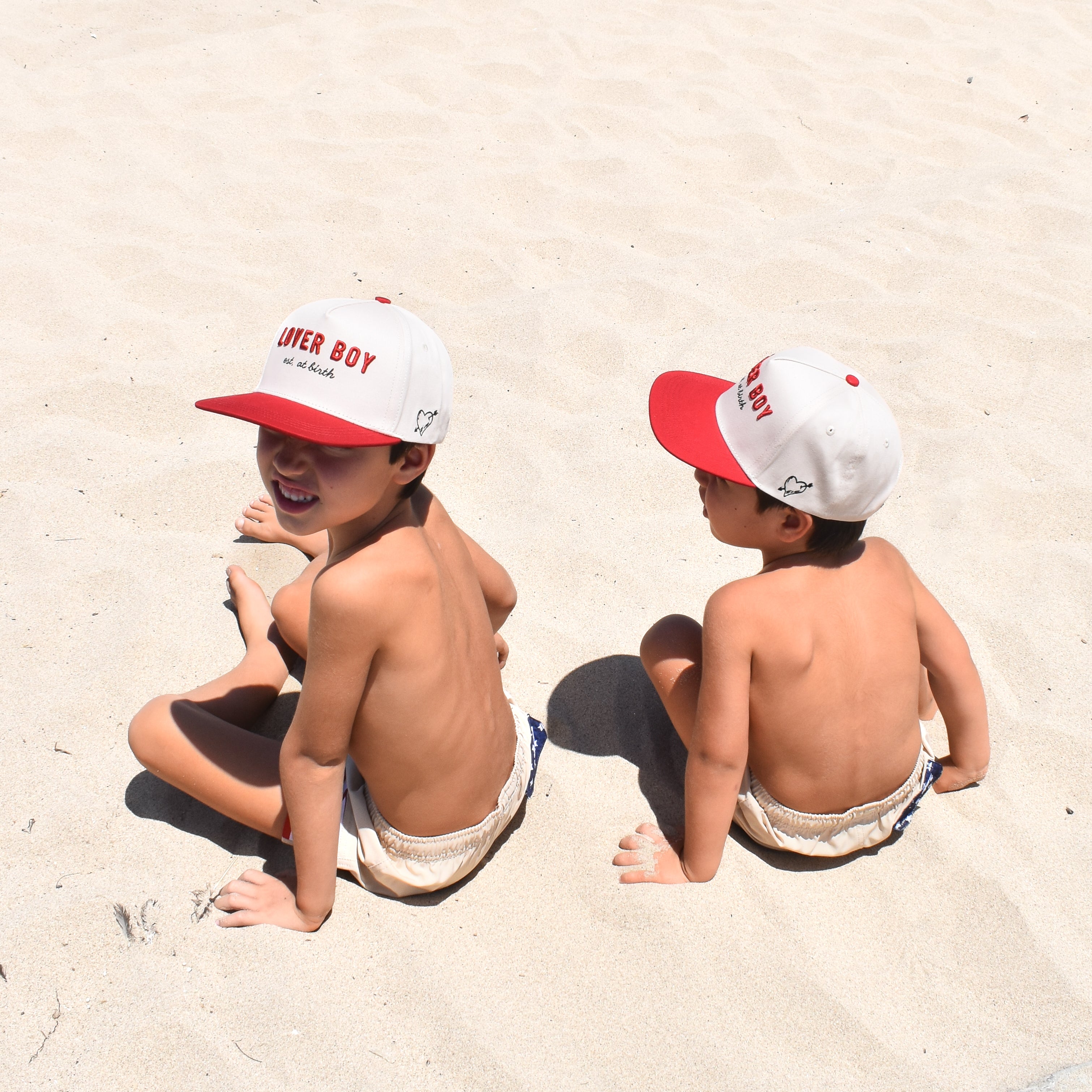 Two children sitting on sand wearing matching red and white hats.