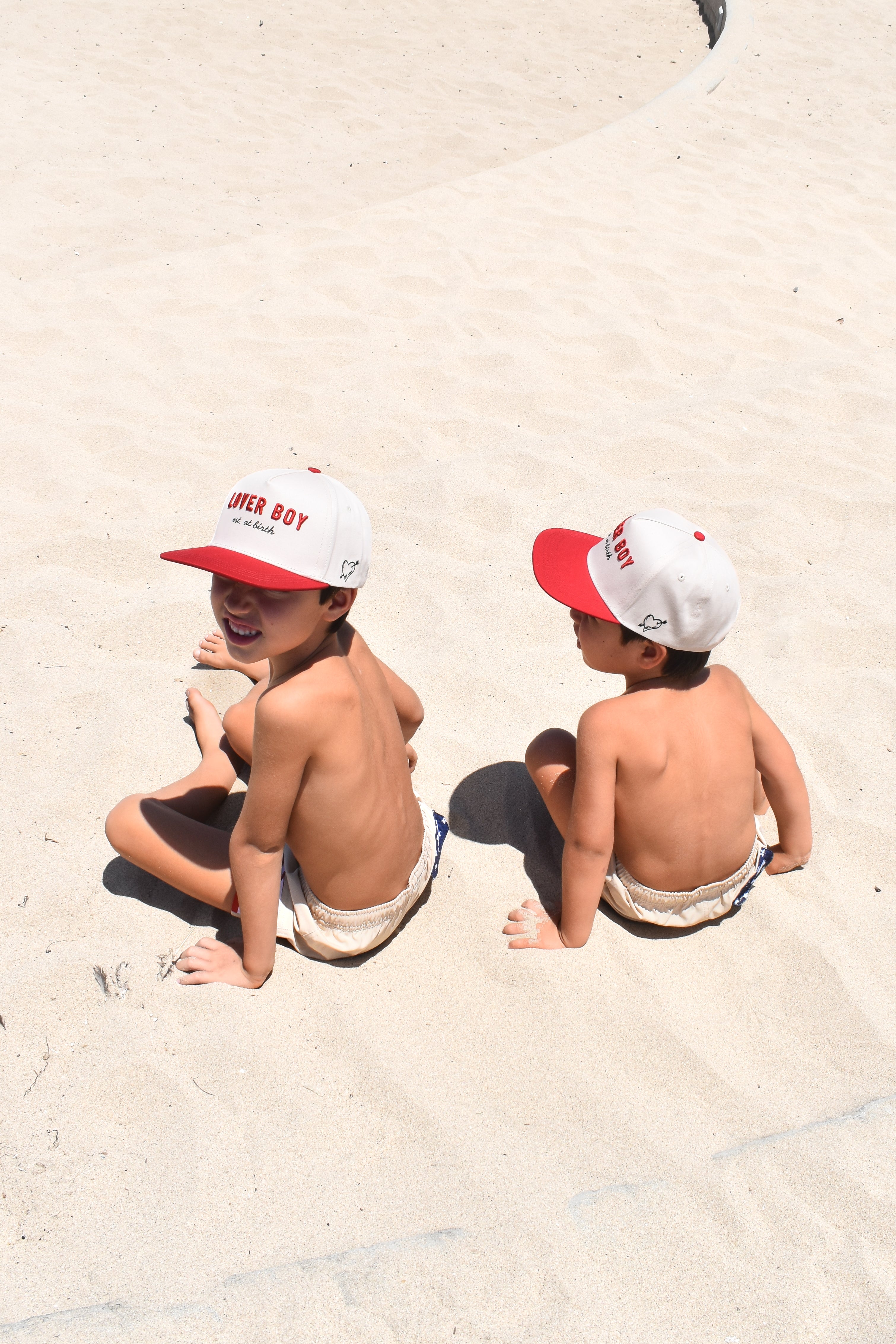 Two children sitting on sand wearing matching red and white hats.