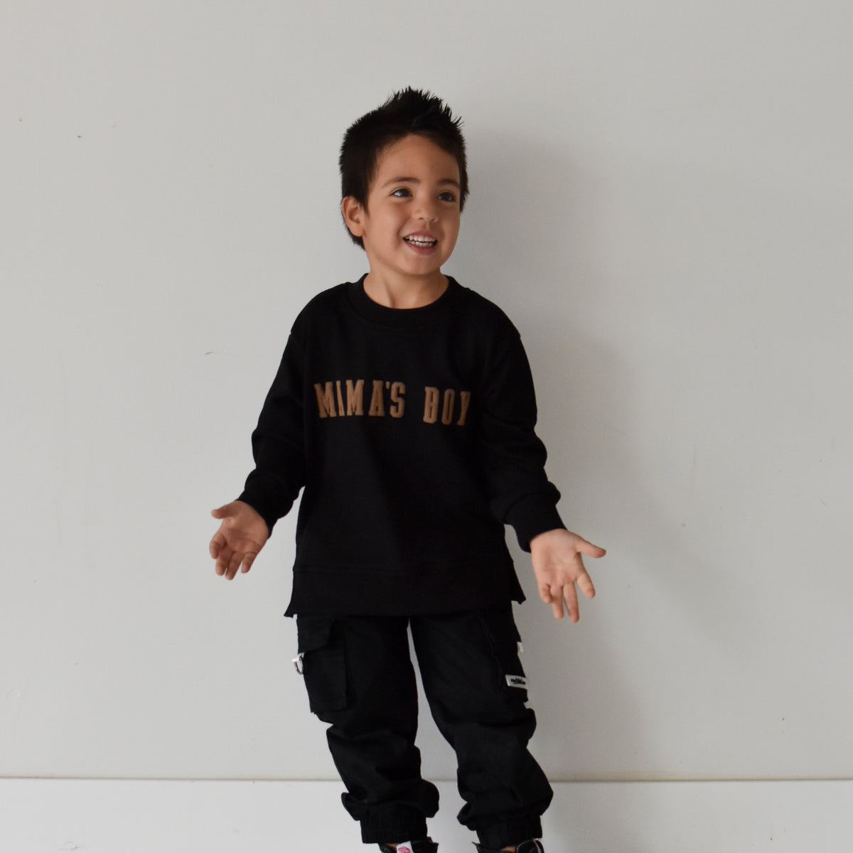 A young boy wearing a black long-sleeve crewneck sweatshirt with 'MAMA'S BOY' printed in gold across the chest, paired with black cargo pants and black sneakers, standing against a plain white wall.