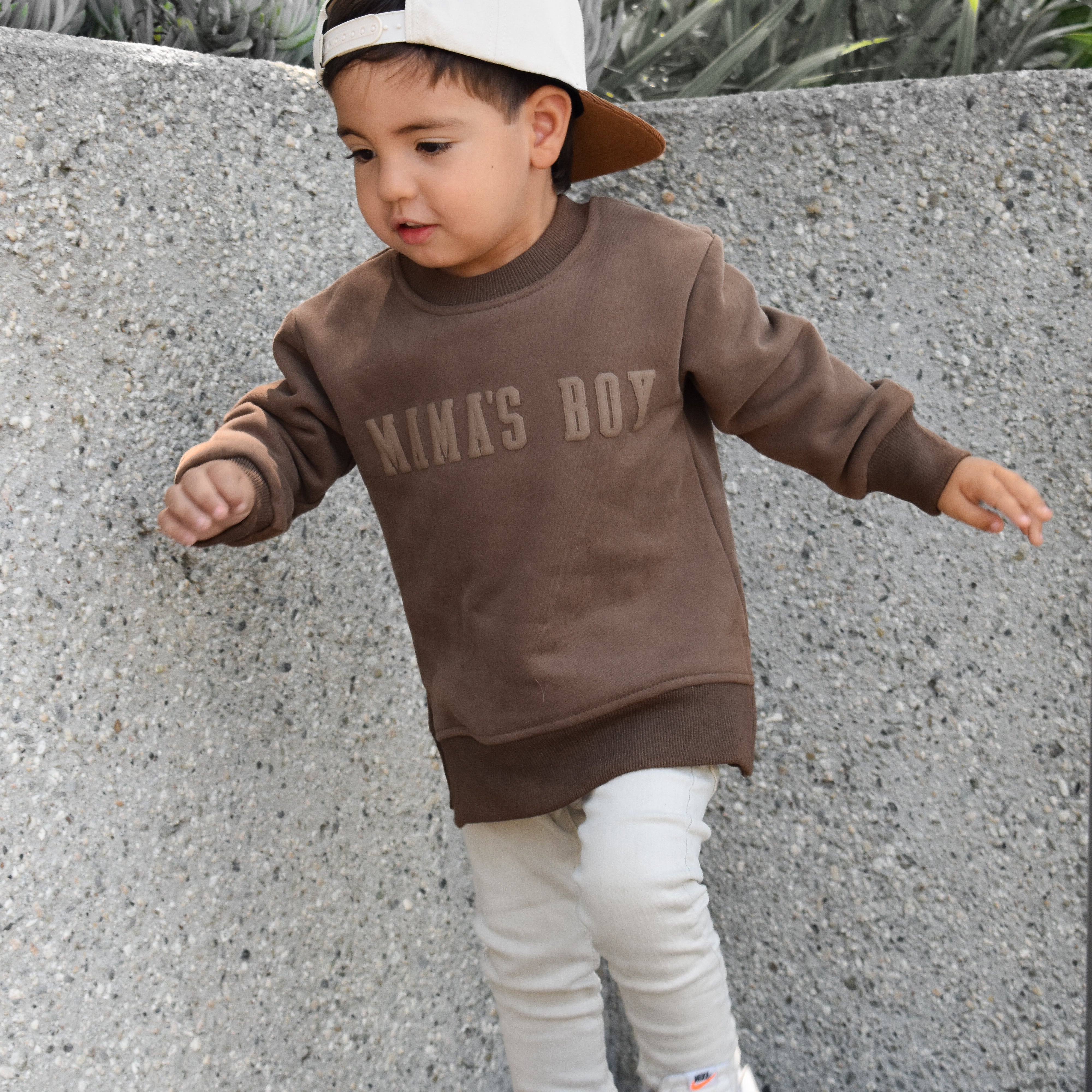 Child wearing a brown 'MamA'S BOY' sweatshirt and white pants standing against a concrete wall.