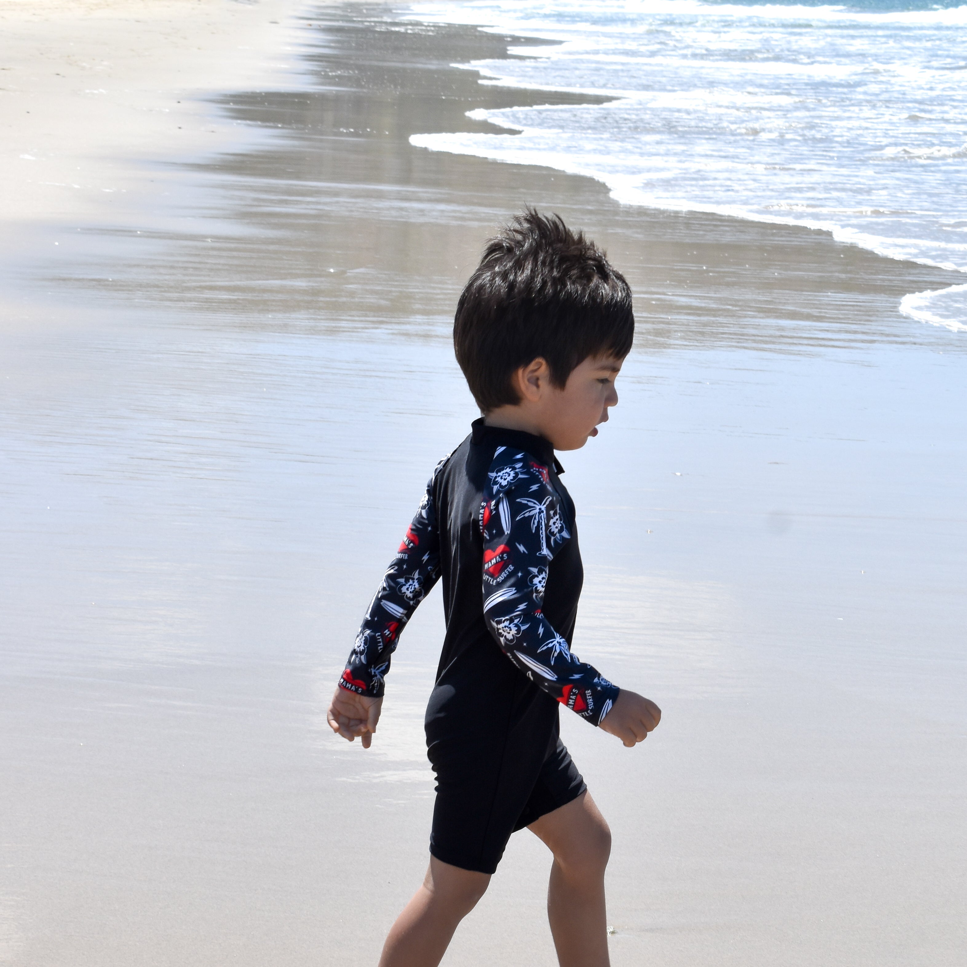 Child in a wetsuit walking on a beach with ocean waves and hills in the background