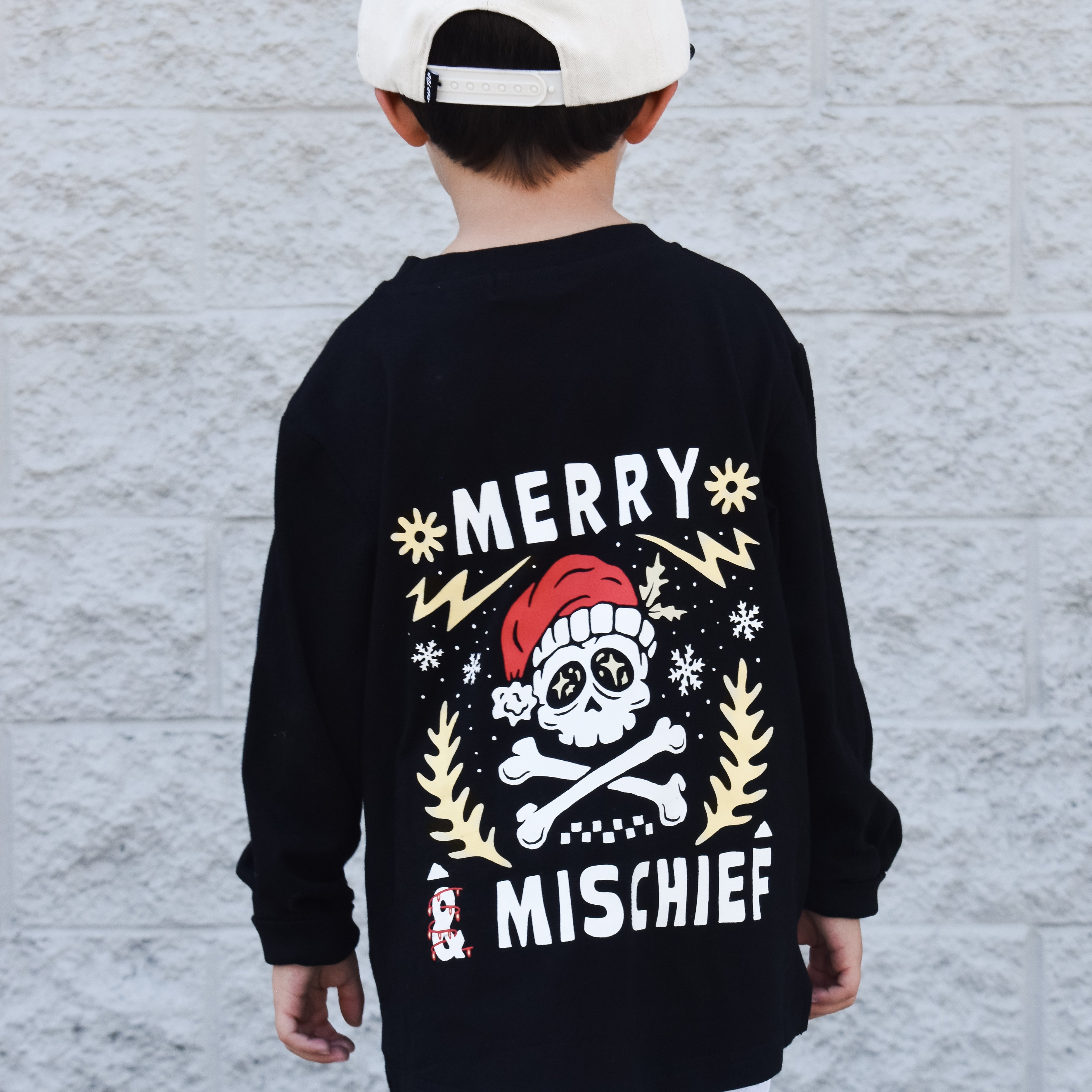 Child wearing a black long-sleeve shirt with a festive design and white cap, standing against a light gray wall.