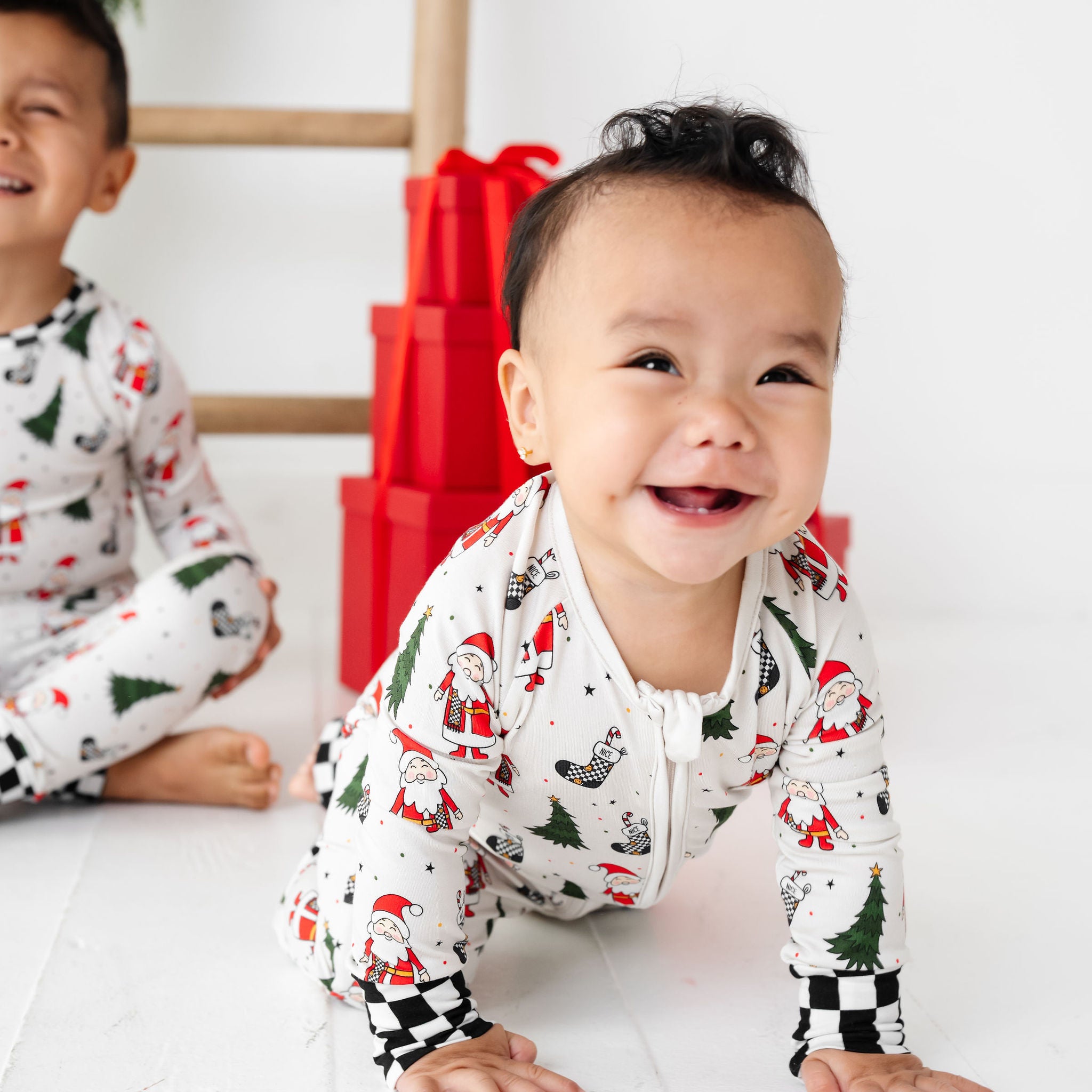 Two children wearing Christmas-themed pajamas on a white floor with red gift boxes in the background.