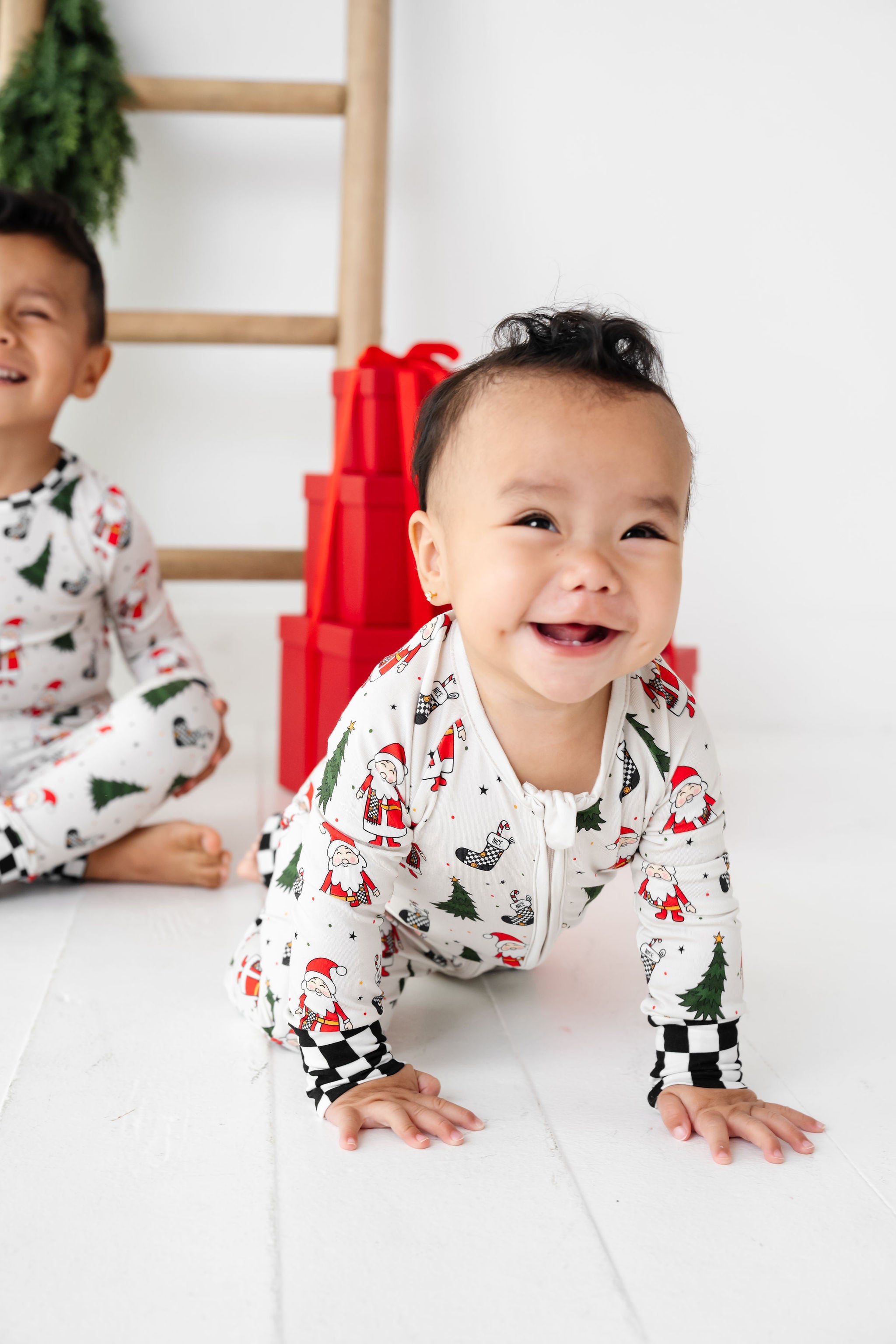 Two children wearing Christmas-themed pajamas on a white floor with red gift boxes in the background.
