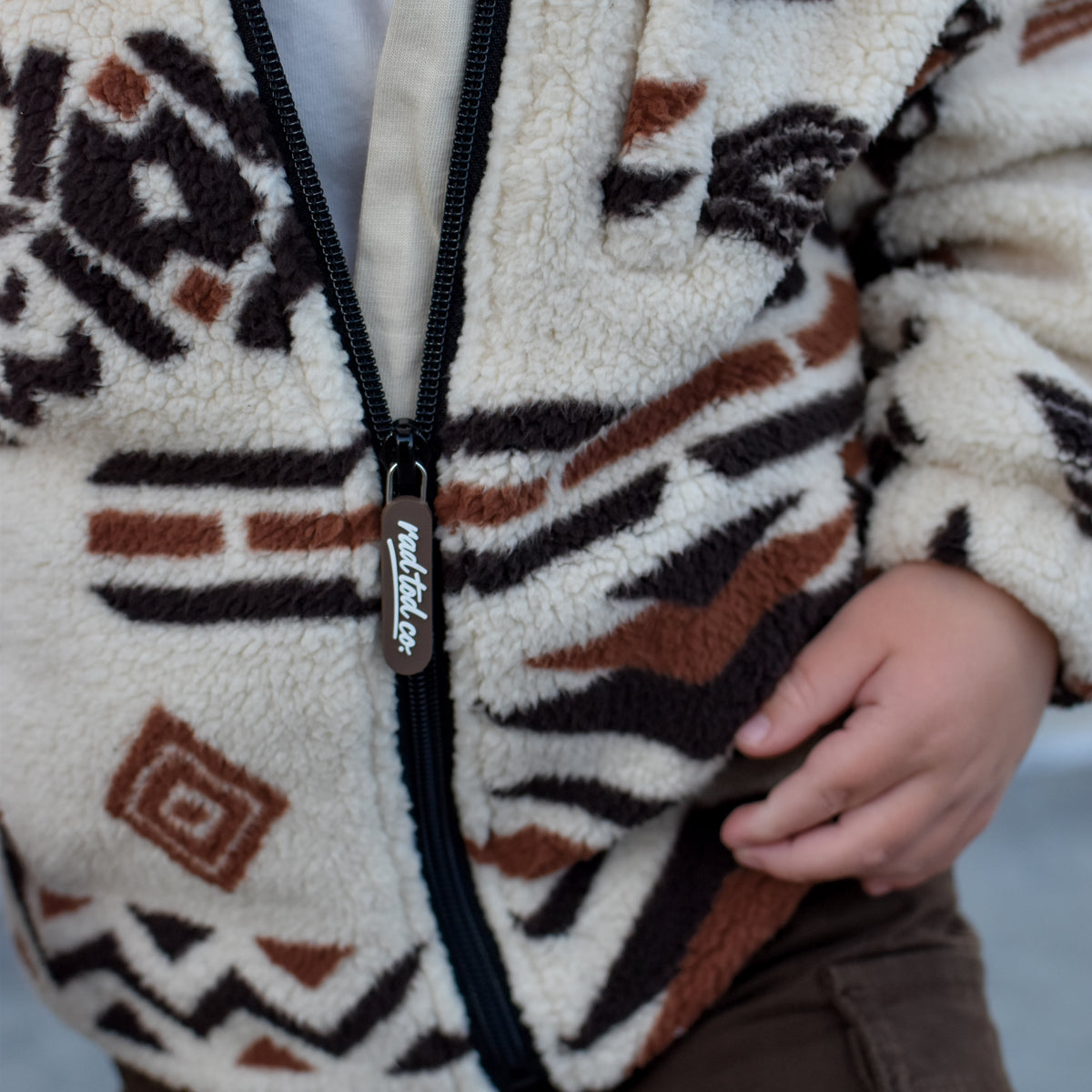 A close-up of a child wearing a sherpa zip-up jacket with a brown, black, and cream geometric pattern, paired with brown cargo pants.
