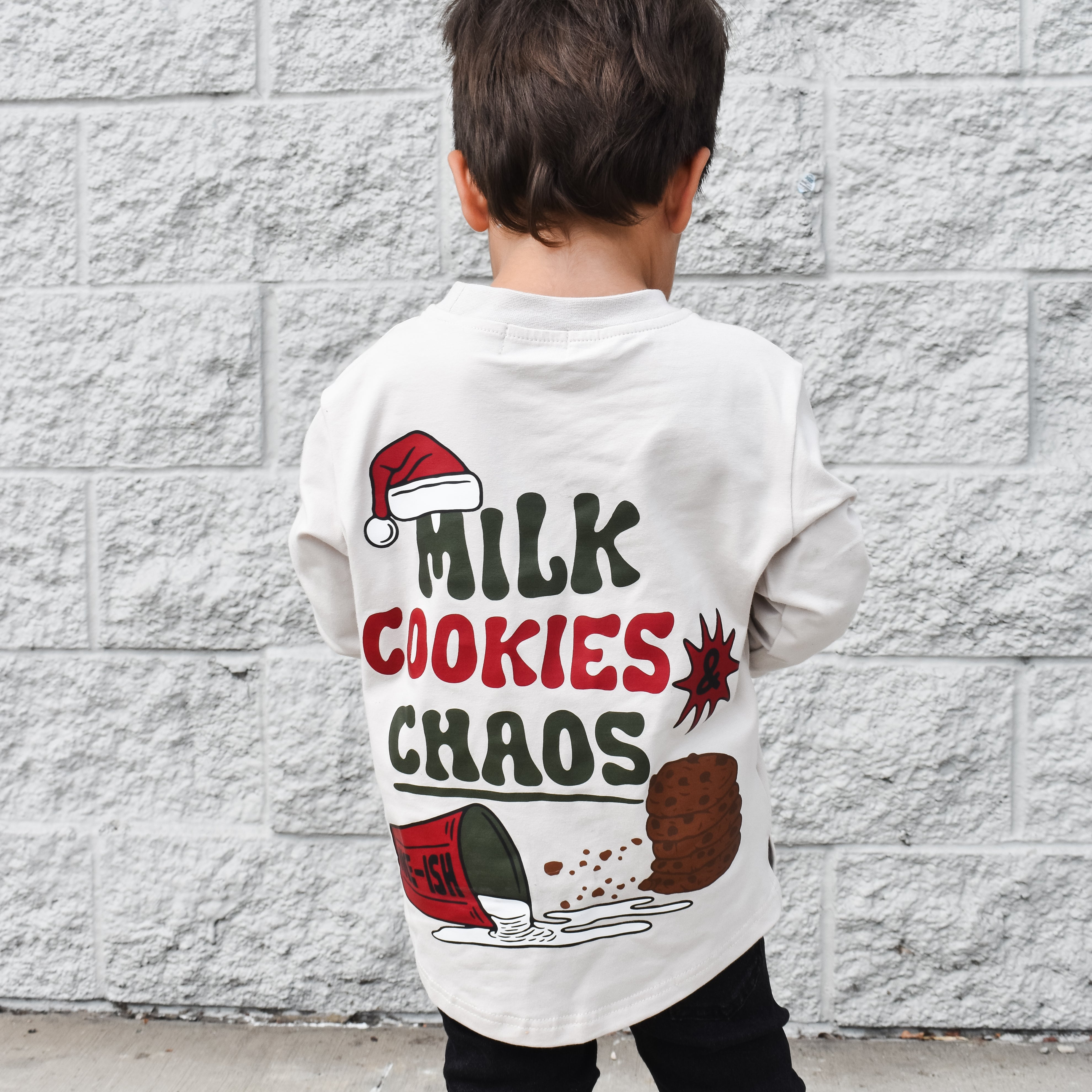 Child wearing a white t-shirt with 'Milk Cookies Chaos' text and graphics against a gray brick wall.