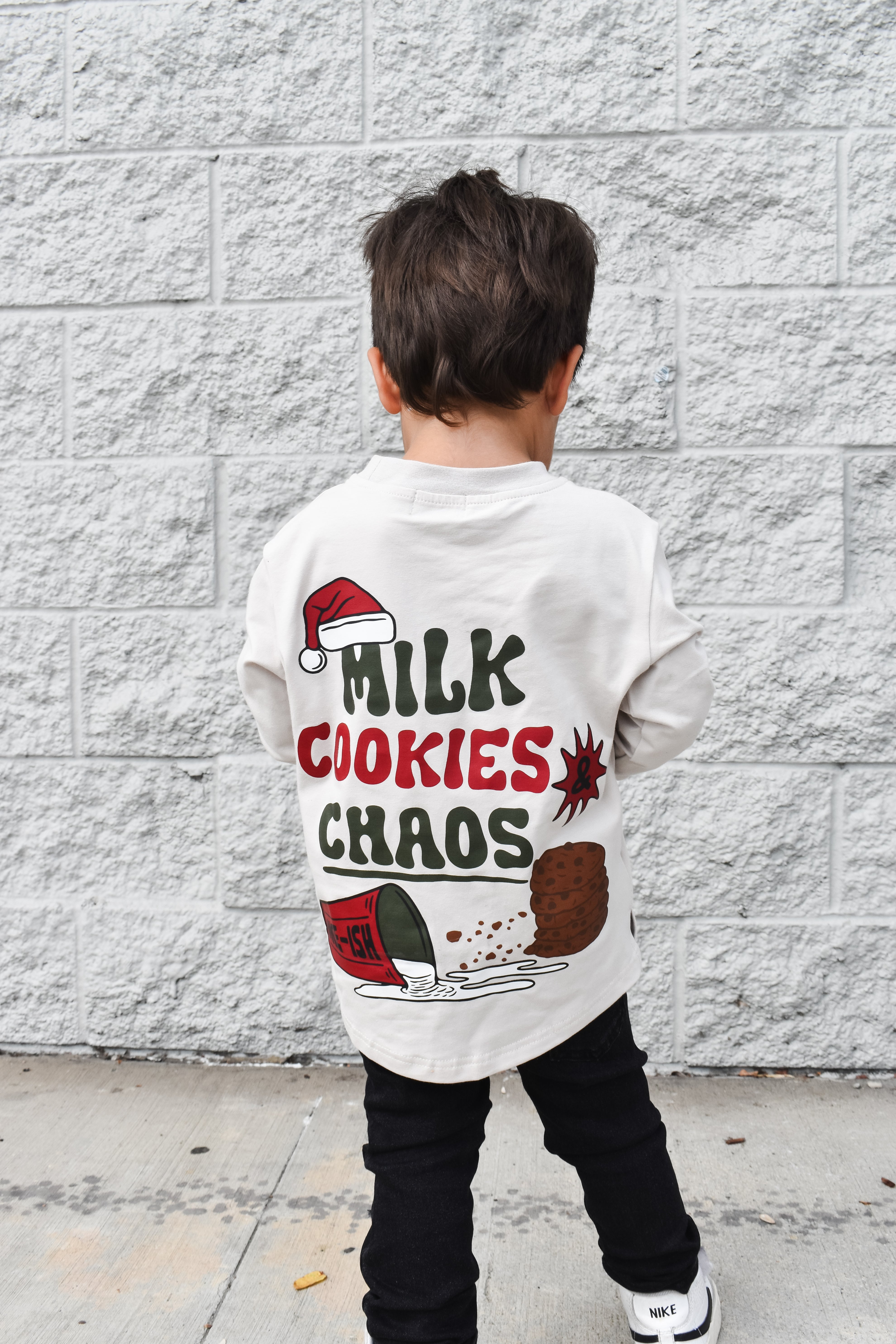 Child wearing a white t-shirt with 'Milk Cookies Chaos' text and graphics against a gray brick wall.