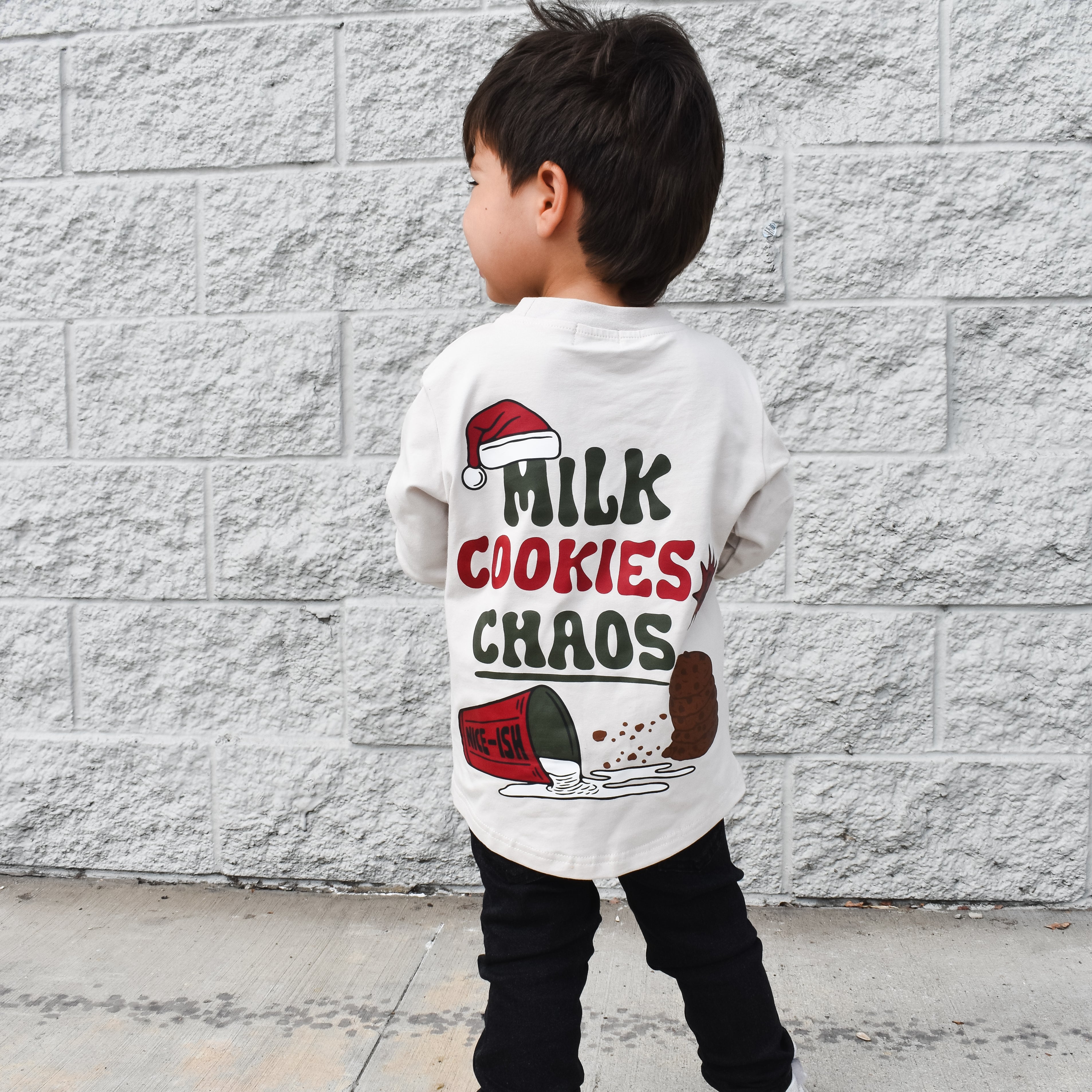 Child wearing a shirt with 'Milk Cookies Chaos' text against a gray brick wall.