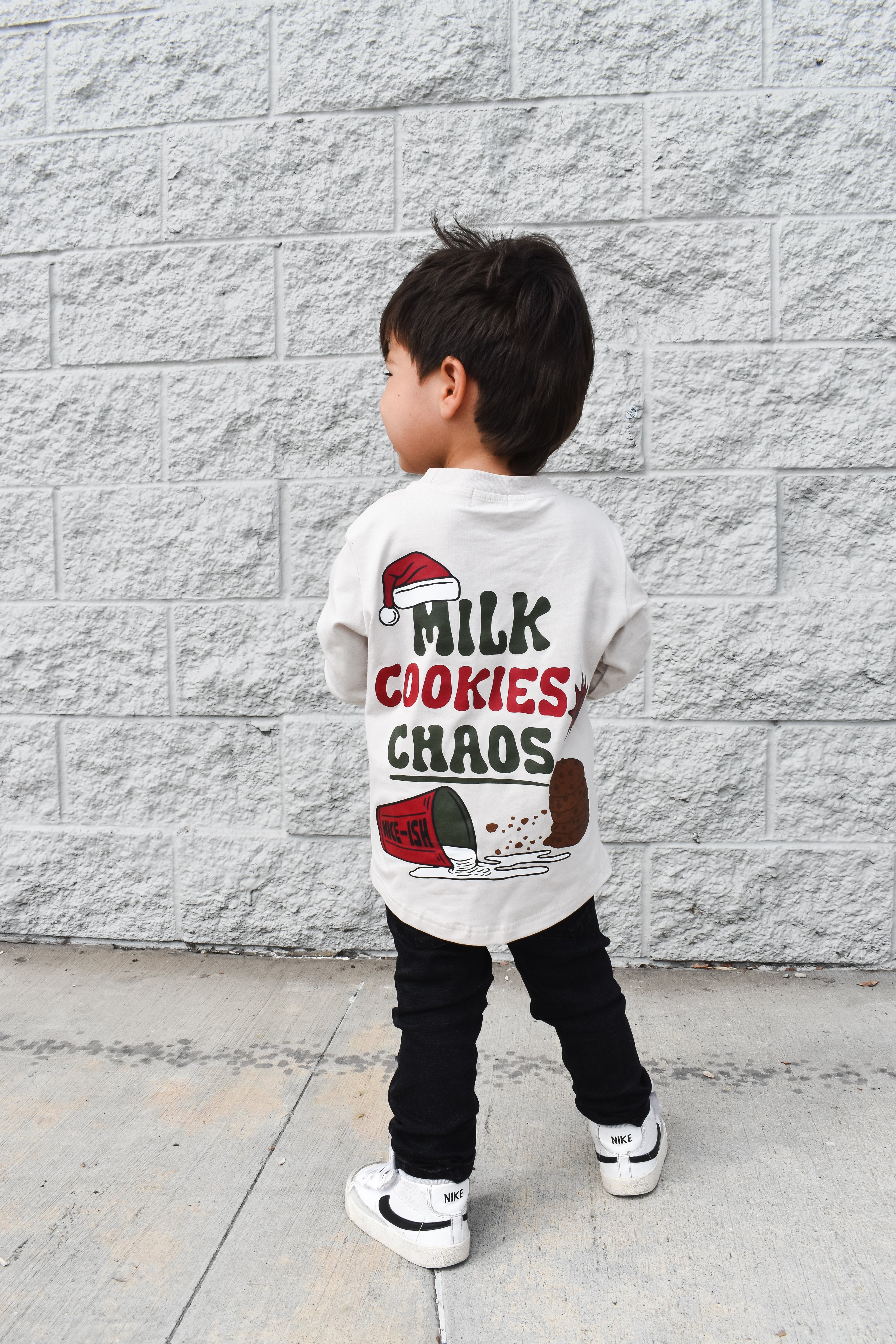 Child wearing a shirt with 'Milk Cookies Chaos' text against a gray brick wall.