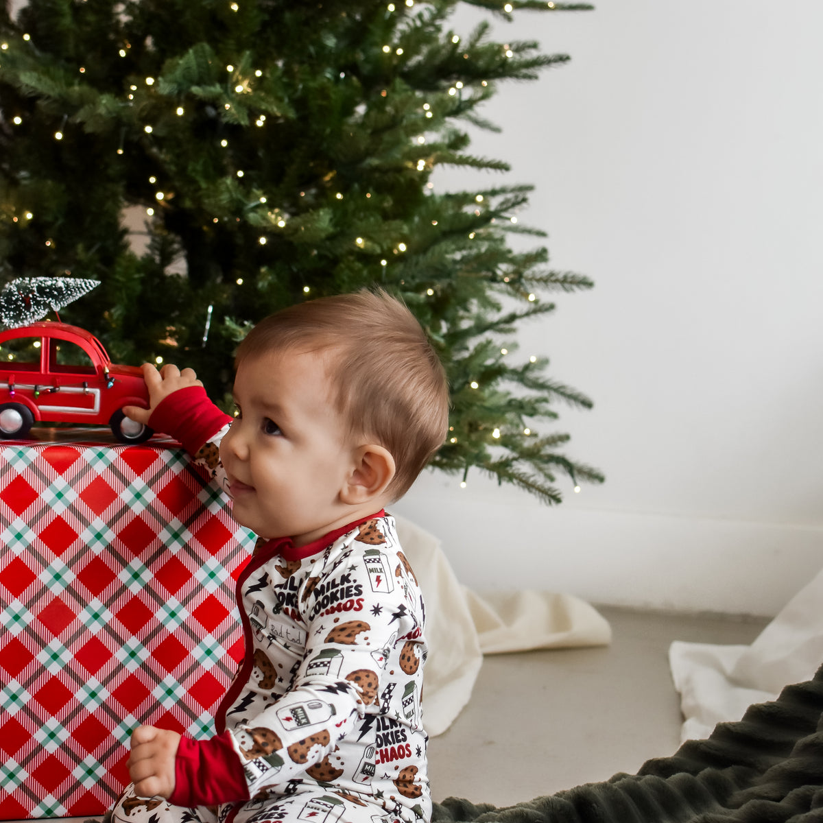 A baby sitting on a dark green textured blanket in front of a Christmas tree, wearing a white long-sleeve pajama set with a brown holiday print and red cuffs. A red and white plaid gift box with a small red toy truck sits to the left.