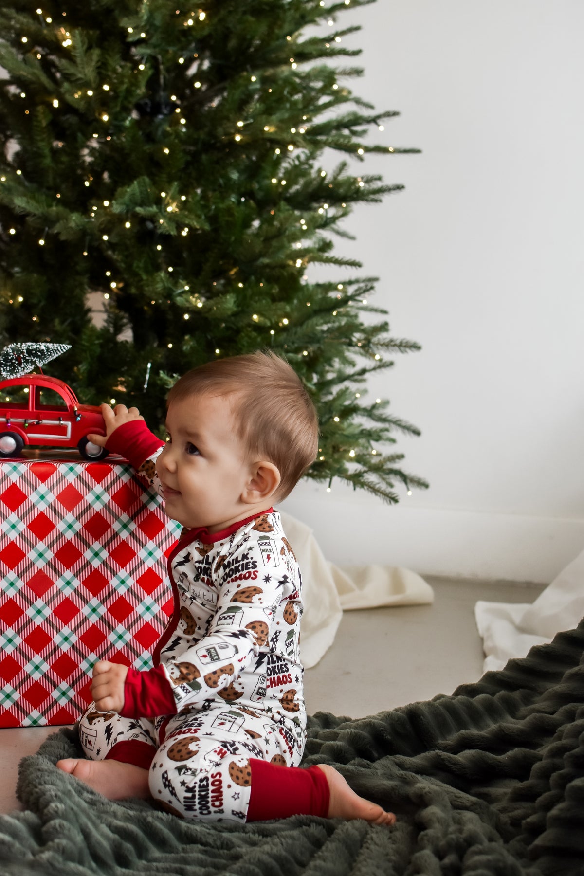A baby sitting on a dark green textured blanket in front of a Christmas tree, wearing a white long-sleeve pajama set with a brown holiday print and red cuffs. A red and white plaid gift box with a small red toy truck sits to the left.