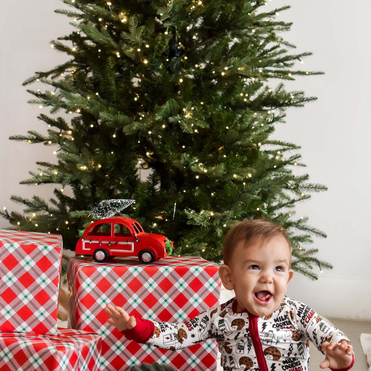 A baby wearing a festive white and red Christmas onesie sits on a gray blanket in front of a decorated Christmas tree, with wrapped presents and a small red toy car nearby.