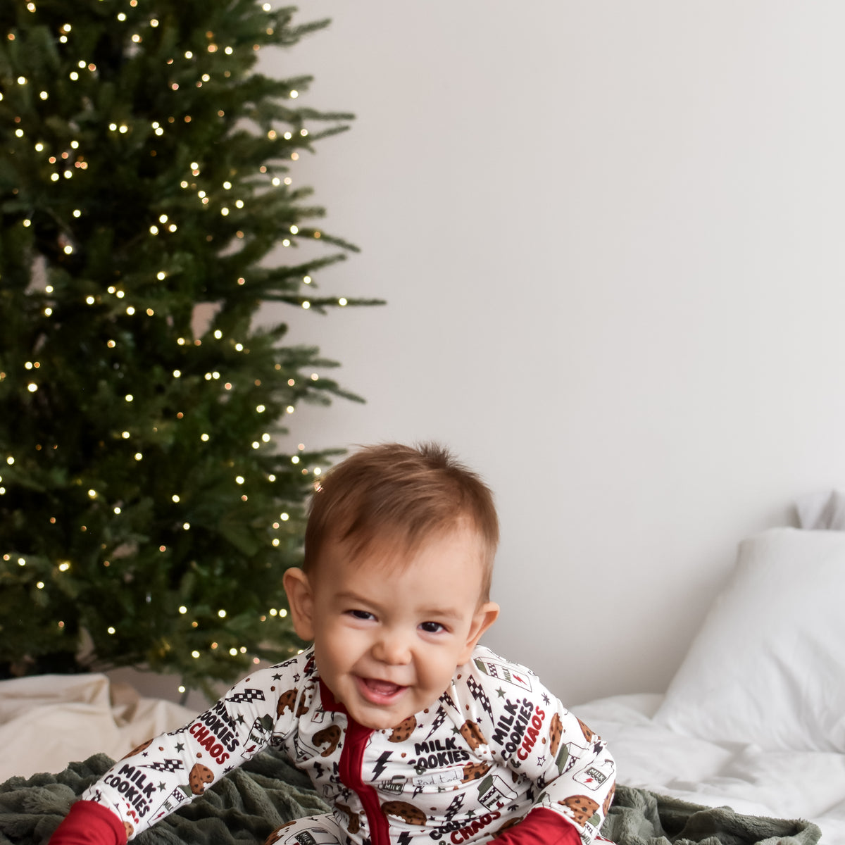 A baby wearing a festive white and red Christmas-themed onesie sits on a cozy green blanket in front of a decorated Christmas tree.