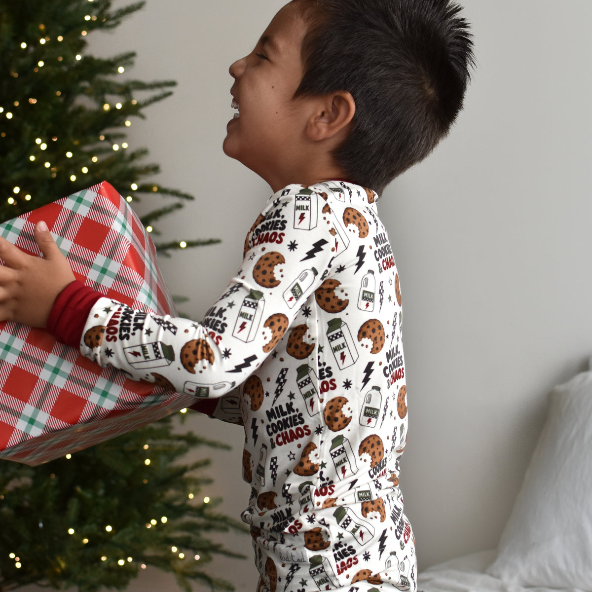 A young boy wearing a white Christmas-themed pajama set with milk cookies and chaos text, sitting on a dark green blanket next to a lit Christmas tree and holding a wrapped gift.