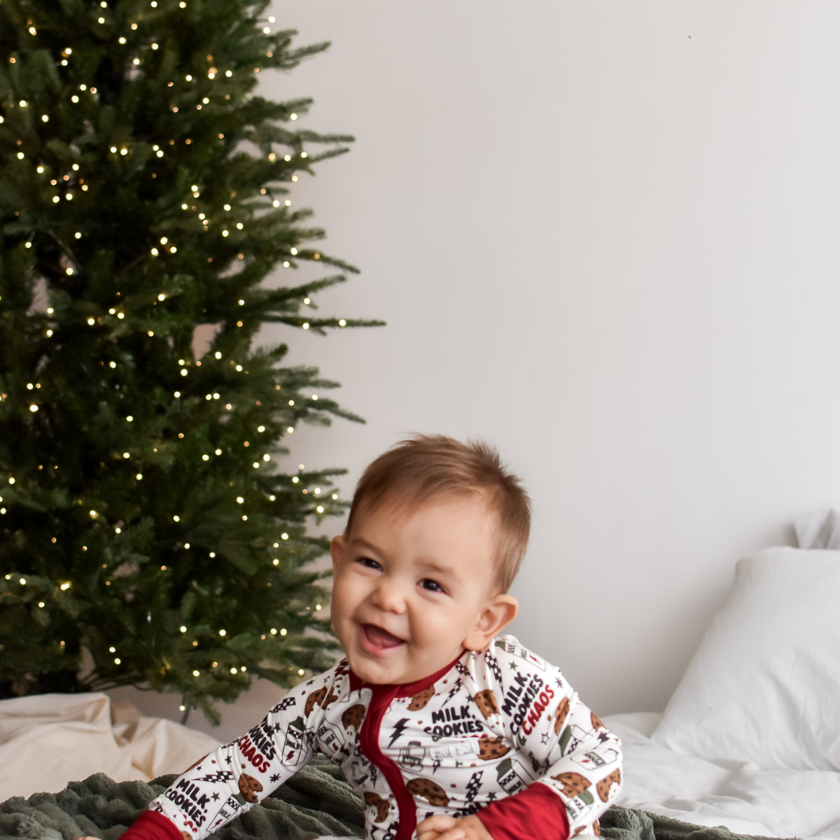 A baby sitting on a bed wearing a festive holiday-themed onesie with a white background, red trim, and a print of gingerbread cookies and reindeer. A decorated Christmas tree and a gray knit blanket are visible in the background.
