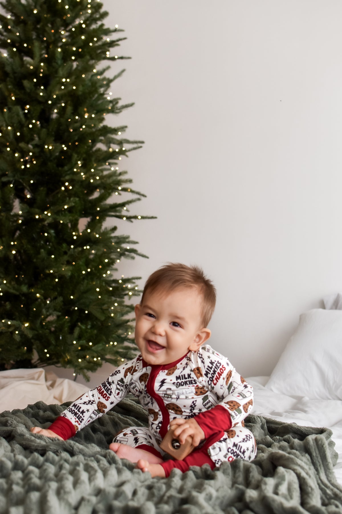 A baby sitting on a bed wearing a festive holiday-themed onesie with a white background, red trim, and a print of gingerbread cookies and reindeer. A decorated Christmas tree and a gray knit blanket are visible in the background.